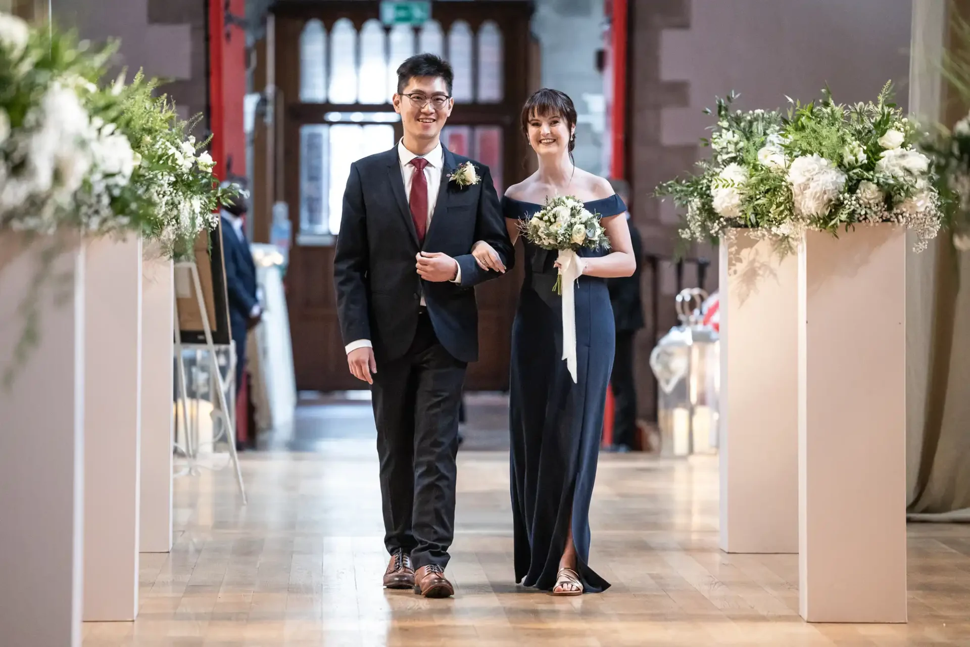 A man in a suit and a woman in a dark gown walk arm in arm down an aisle decorated with flowers.