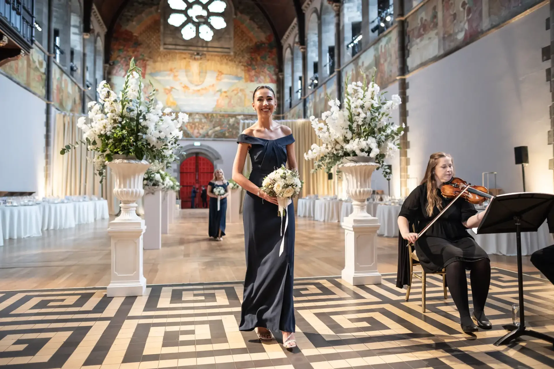 A woman in a dark gown walks down an ornate aisle holding a bouquet. A seated musician plays a string instrument. Large floral arrangements flank the aisle in an elegant hall.