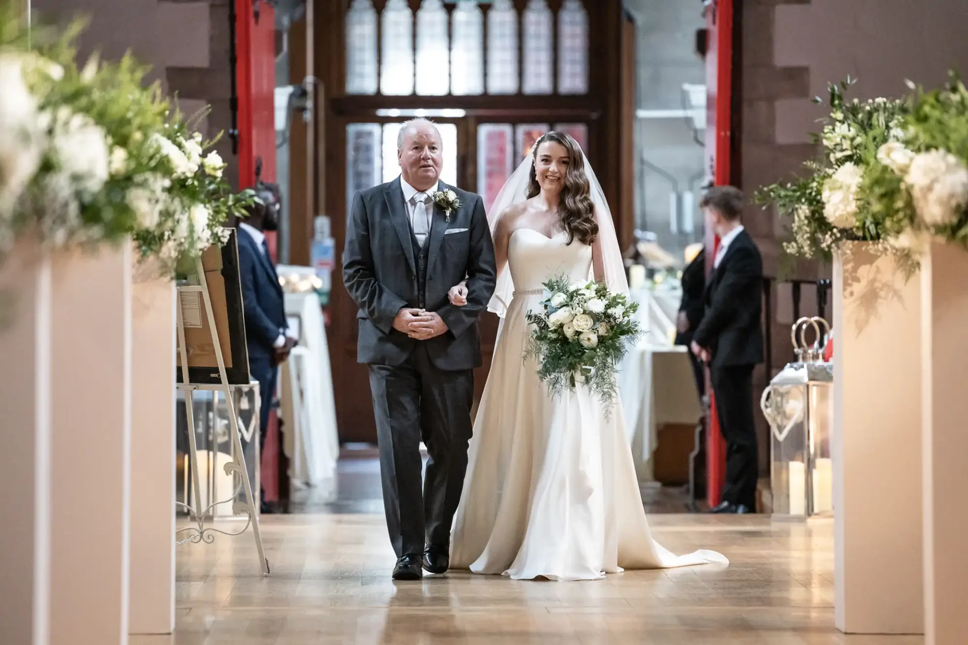 A bride in a white gown walks down an aisle with a man in a suit. She holds a bouquet, and both are smiling. Floral arrangements line the walkway.