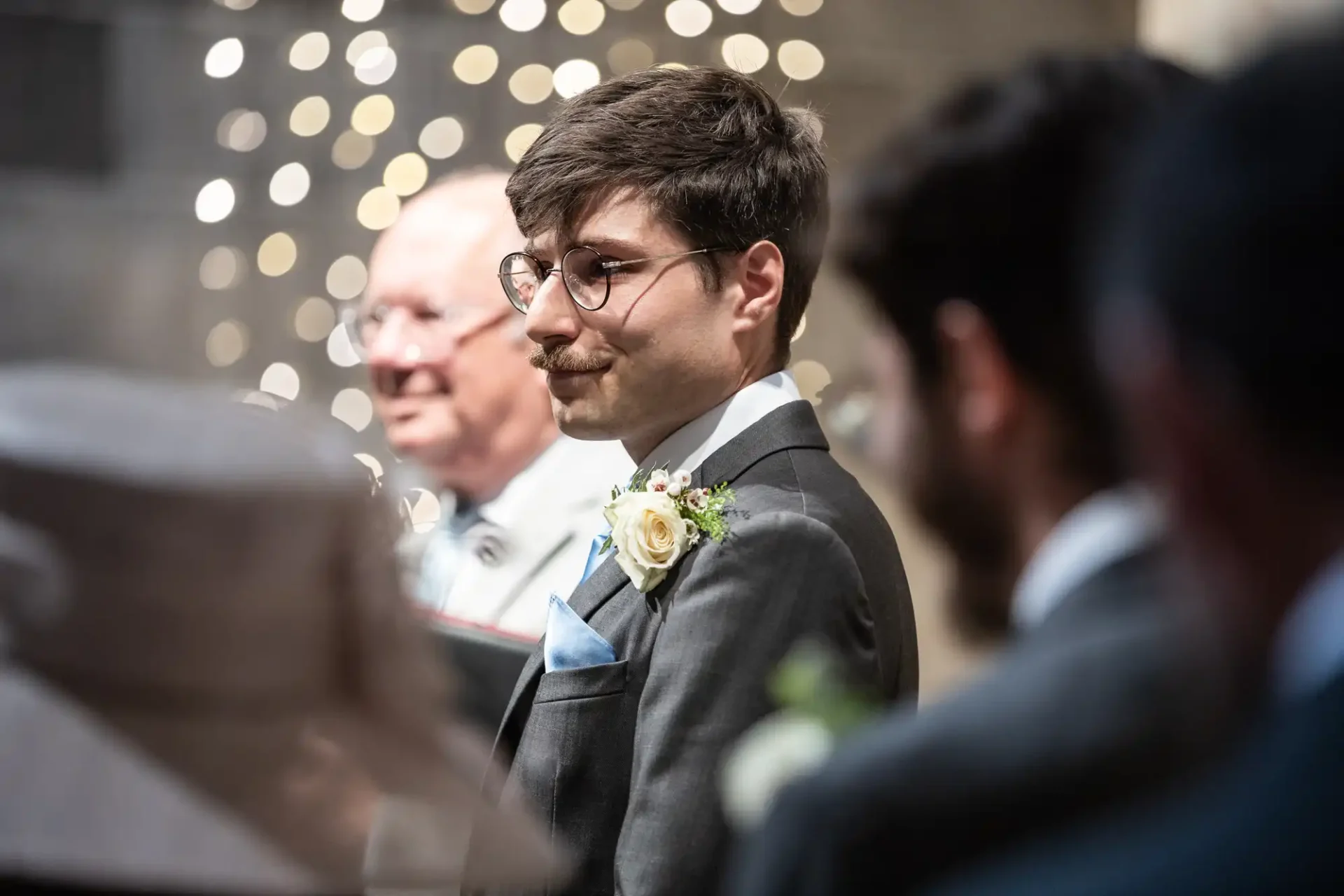 A man in a suit with a boutonniere stands among others at a formal event, possibly a wedding, with soft lights in the background.