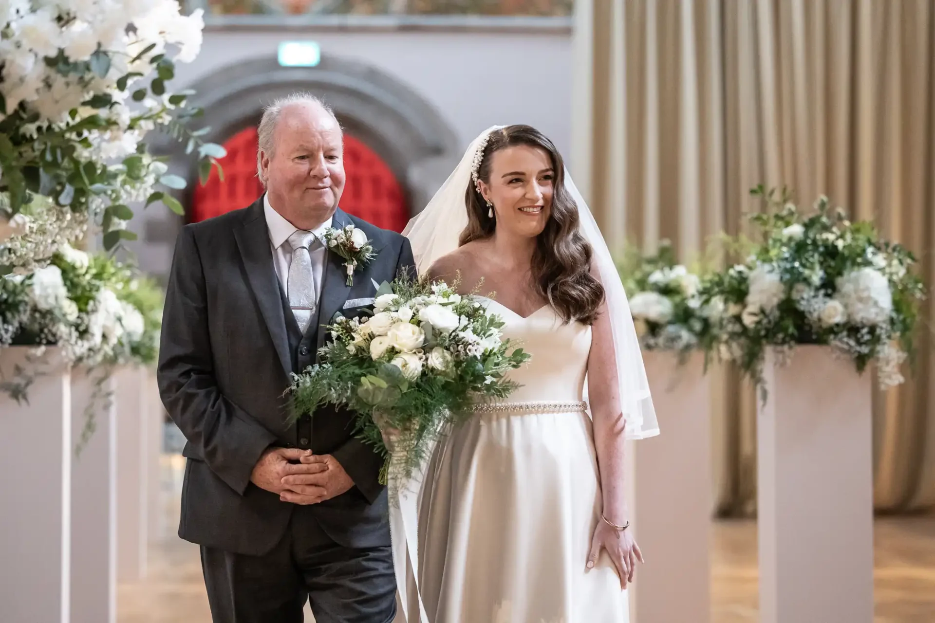 A bride in a white dress and veil walks down the aisle with an older man in a suit, surrounded by white flowers and greenery.