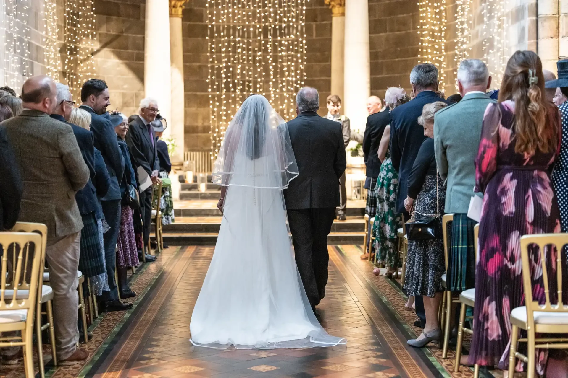 A bride walks down the aisle with an older man in a church filled with seated guests.