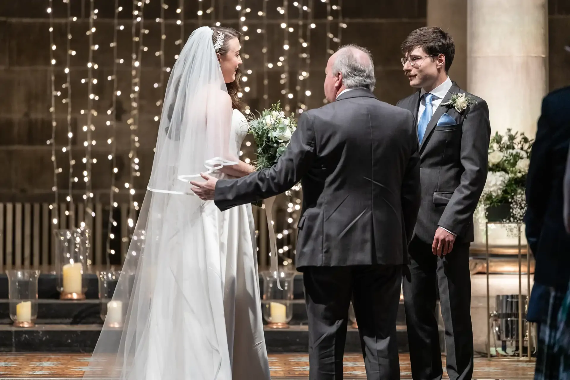 A bride and groom stand in front of an older man during a wedding ceremony in a dimly lit venue with string lights and candles.