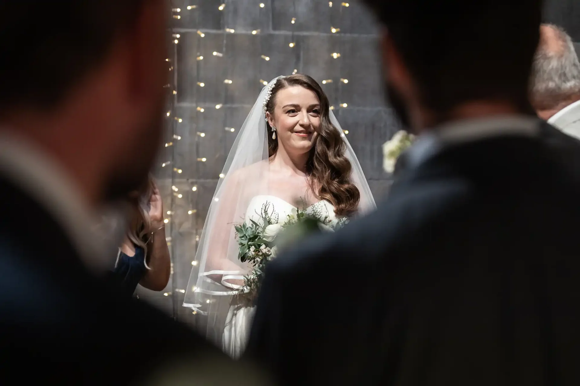 Bride in a white dress holds a bouquet, standing under string lights, framed by blurred silhouettes of people at a wedding ceremony.