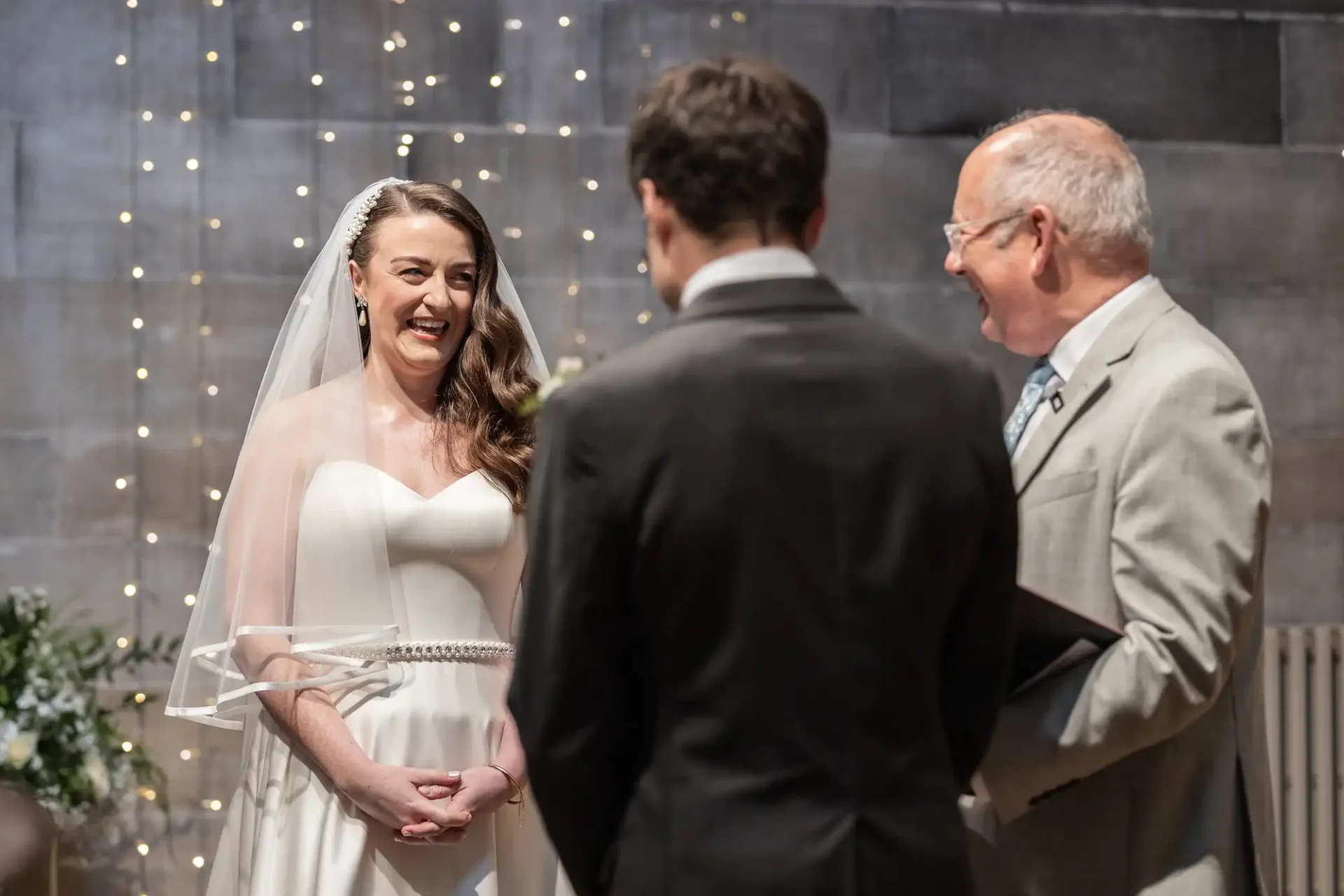 A bride in a white dress stands smiling, facing a groom and officiant. They are in a decorated indoor setting with string lights.