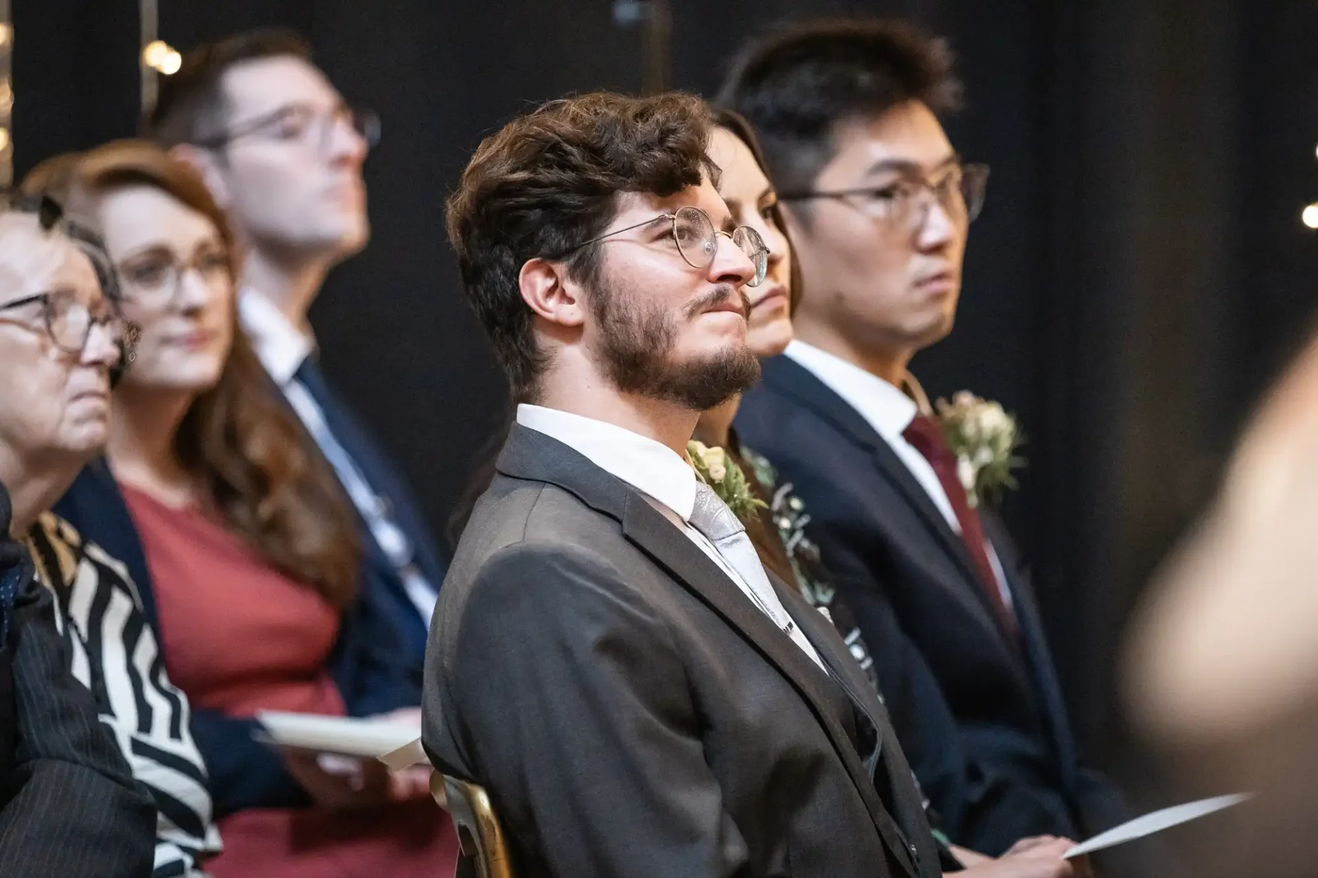 A group of seated people in formal attire, attentively watching an event. A man with glasses is in the foreground.