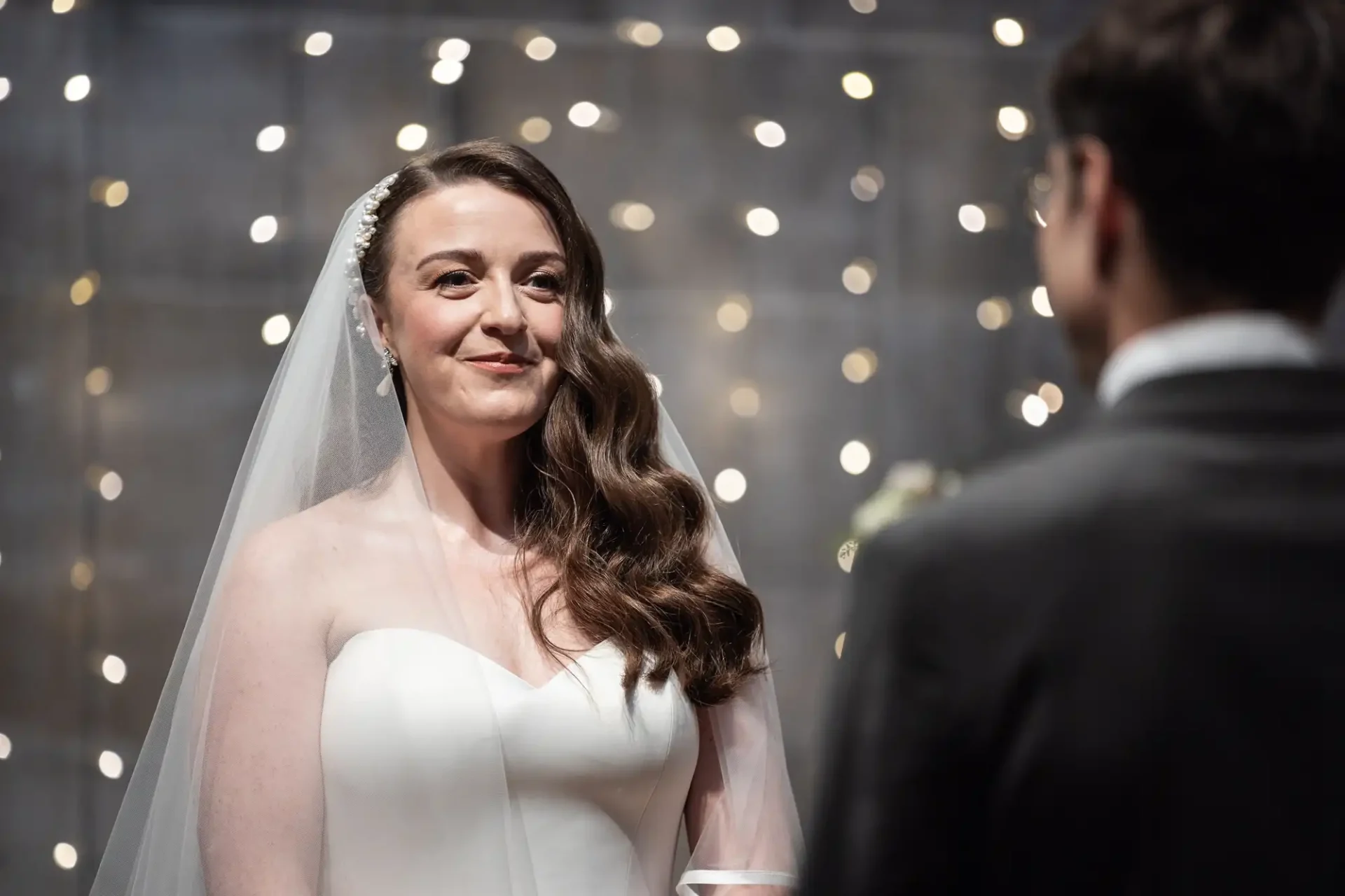 Bride in white gown with a veil stands facing a man in a suit against a backdrop of glowing lights.