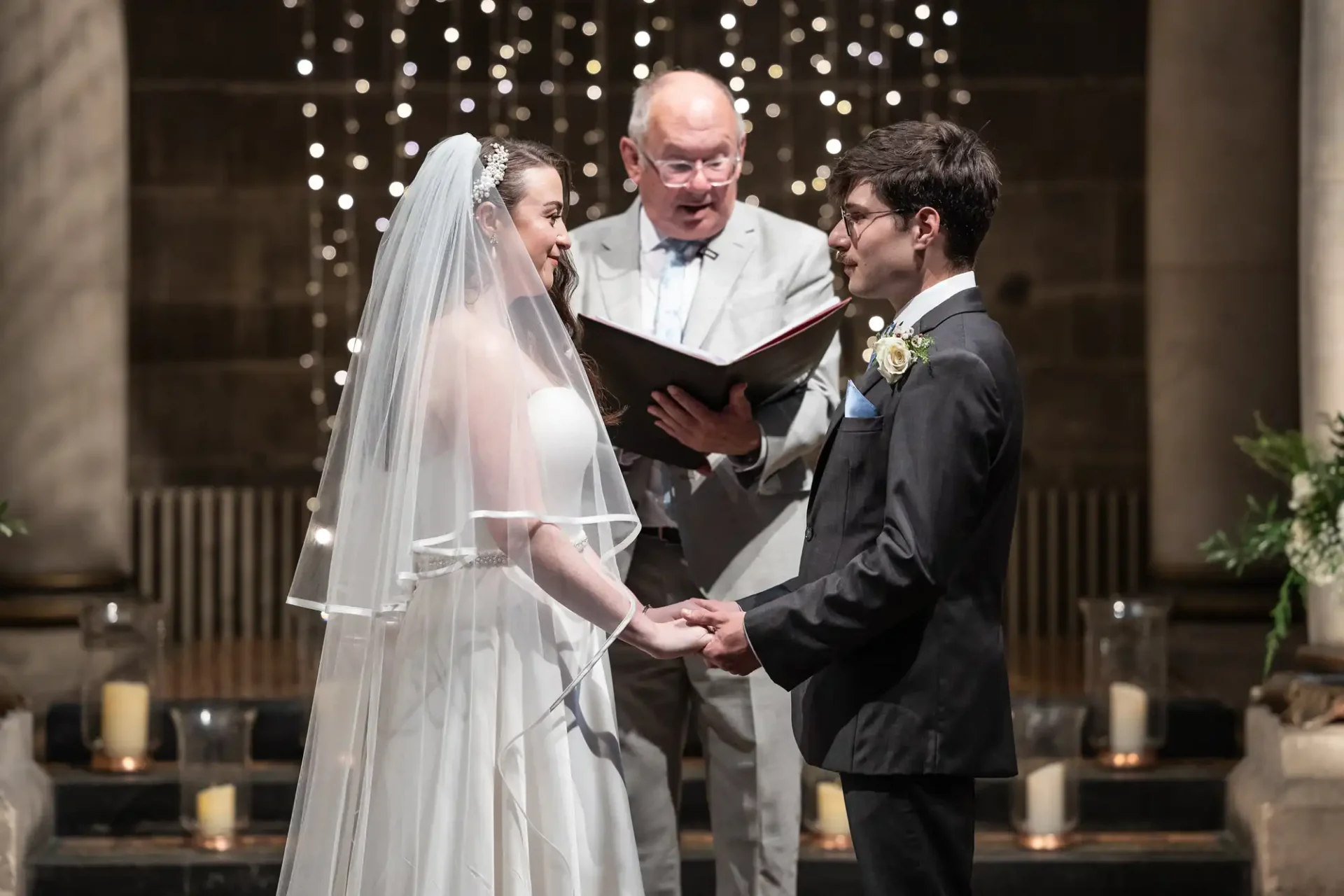 A bride and groom stand holding hands in front of an officiant during a wedding ceremony. The background features string lights and candles.