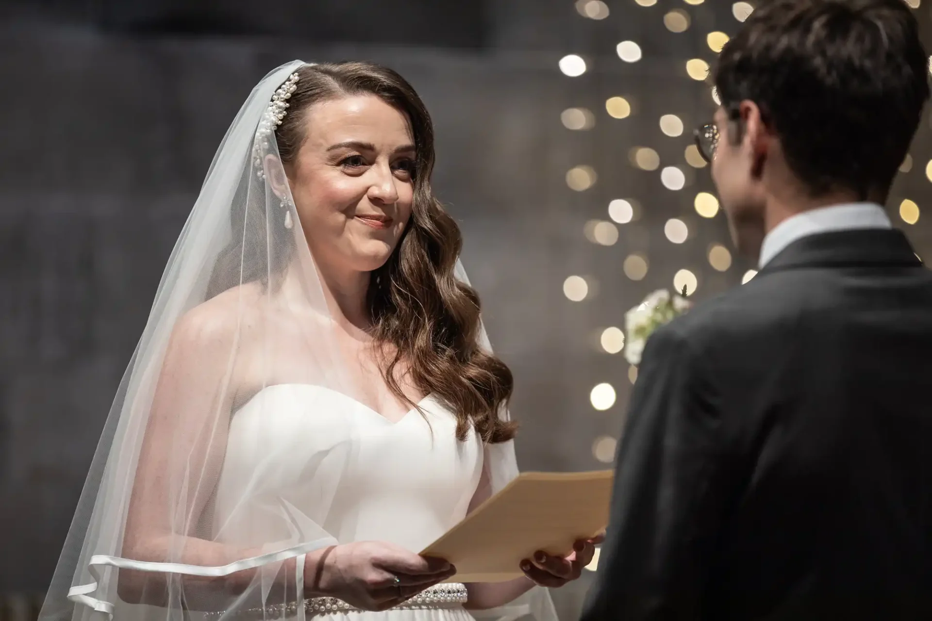 A bride in a wedding dress holds a paper, standing next to a groom in a suit during a wedding ceremony. Blurred lights are in the background.