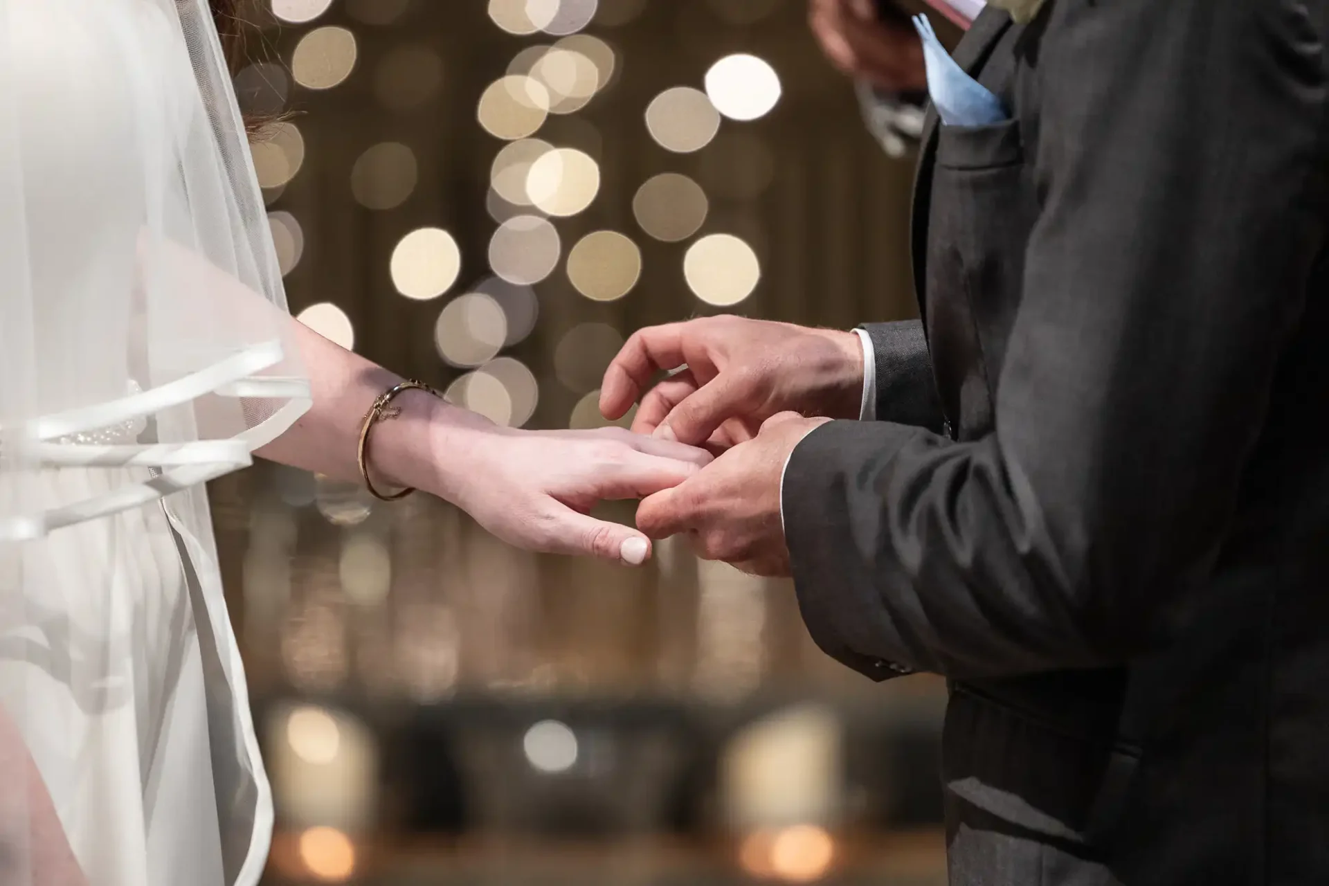 A bride and groom exchange rings during a wedding ceremony, with soft focus lights in the background.