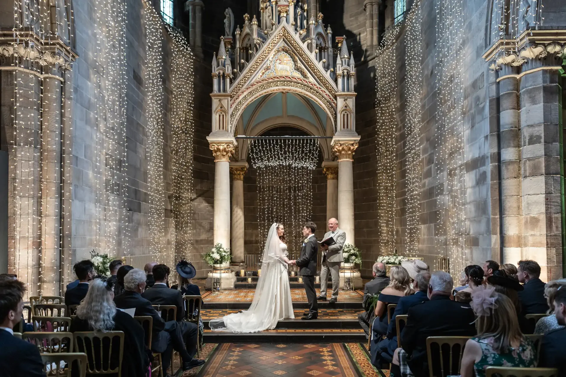 A couple stands at the altar during their wedding ceremony in an ornately decorated church, surrounded by guests seated on either side.