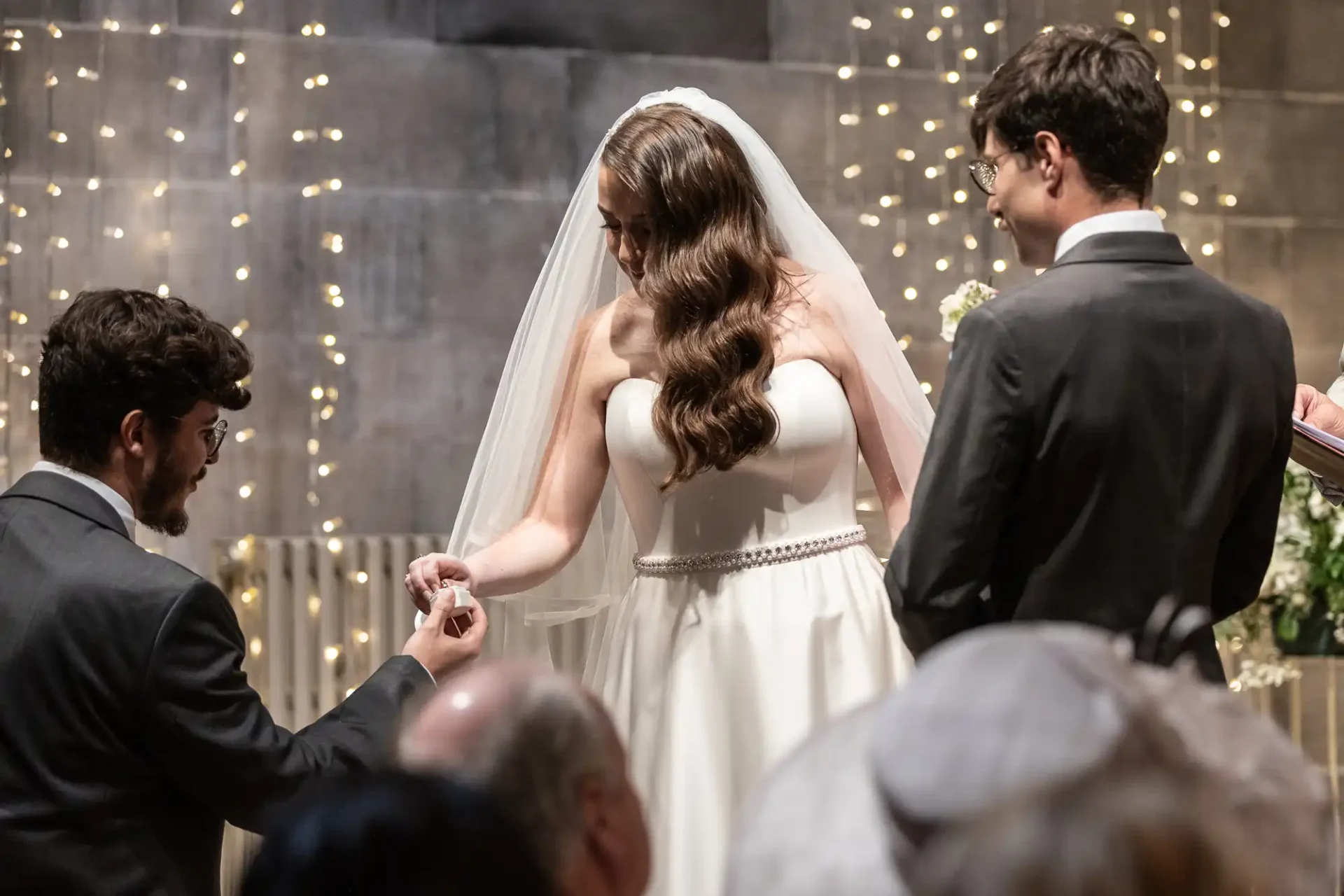 Bride and groom stand together during wedding ceremony, as a man offers them a ring. Fairy lights and flowers decorate the background.