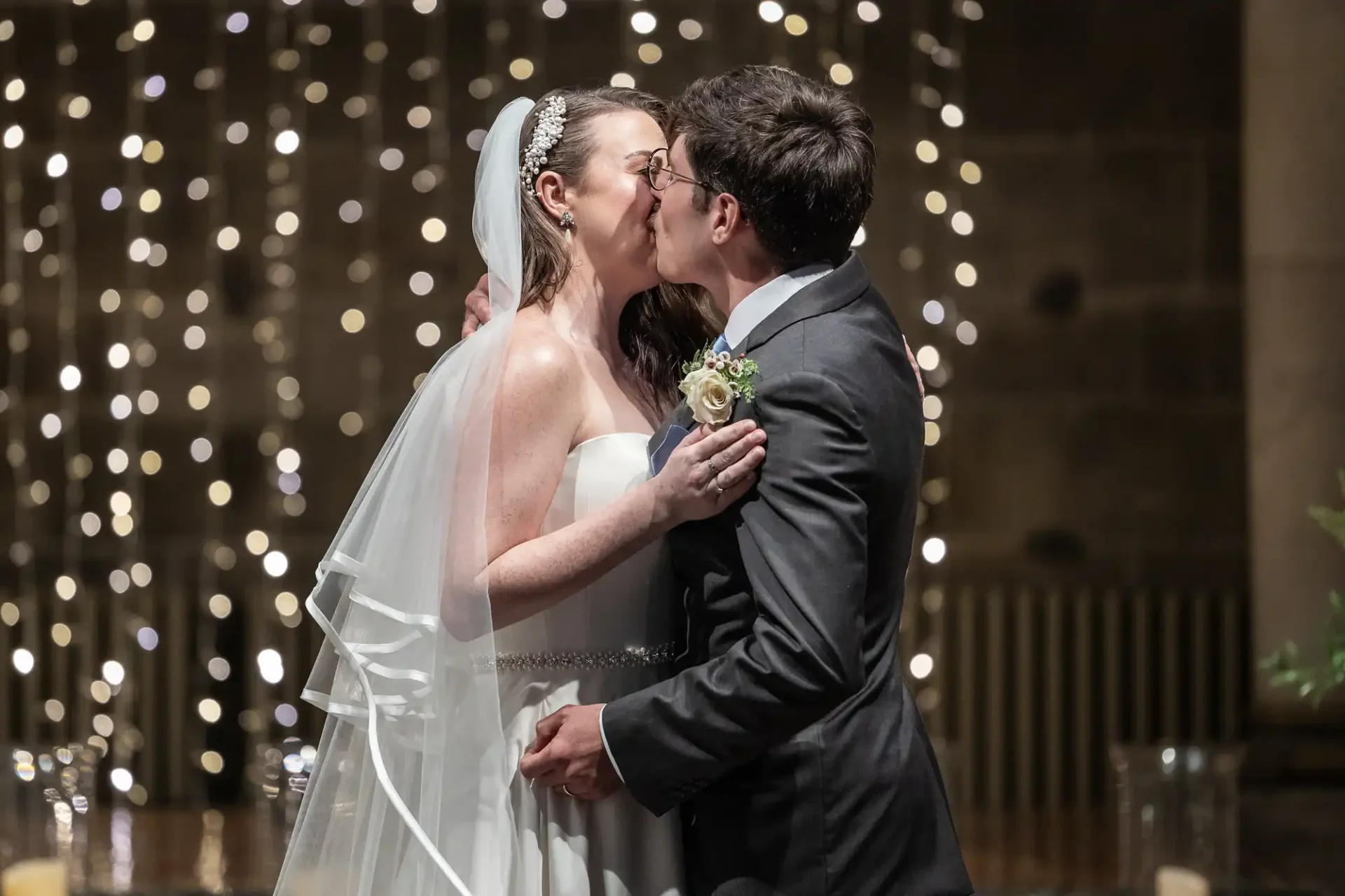 A bride and groom share a kiss at their wedding ceremony. The bride wears a white dress and veil, holding the groom's face. The background features blurred string lights.