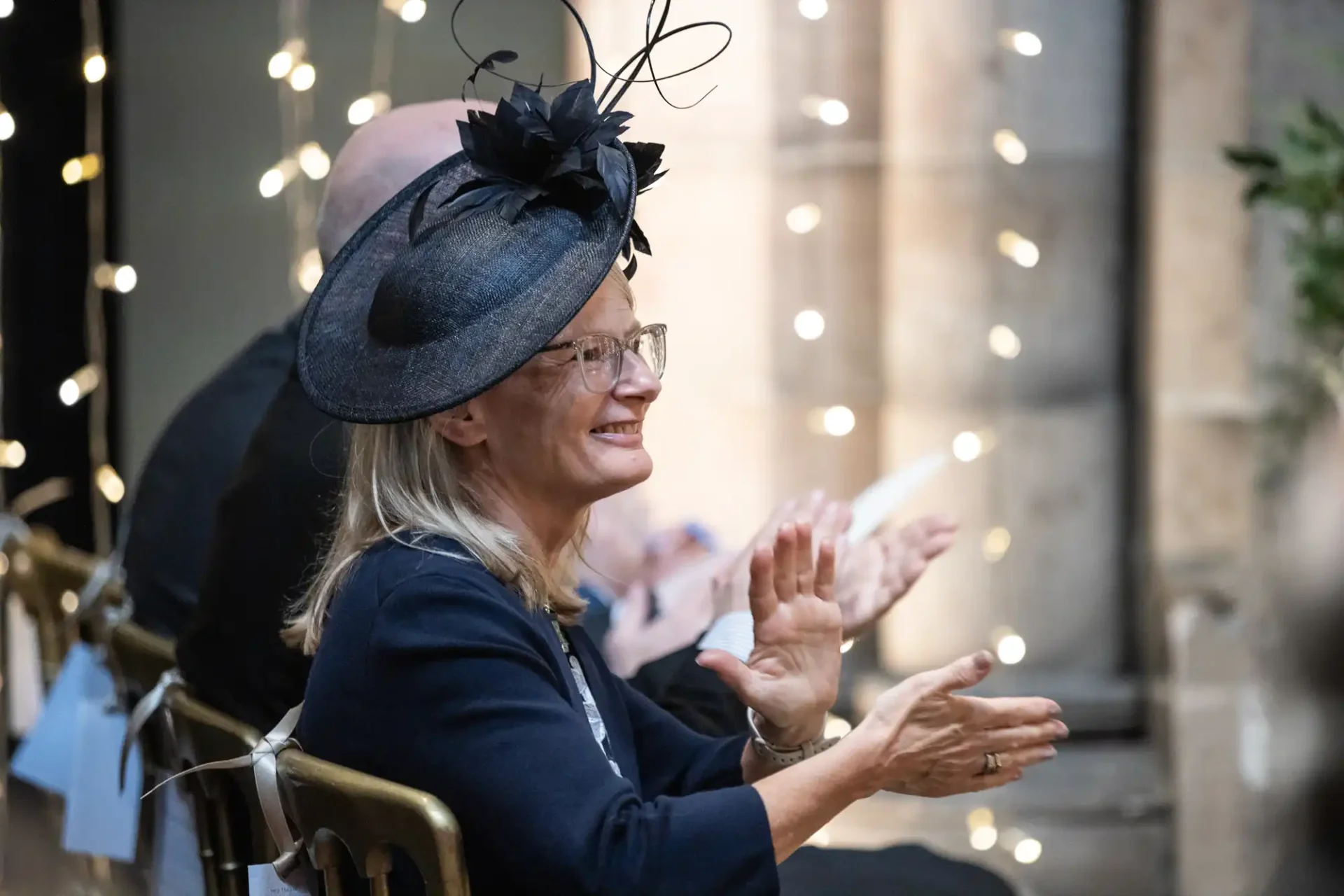 A woman in glasses, wearing a dark hat with decorative feathers, sits and claps in a setting adorned with string lights.