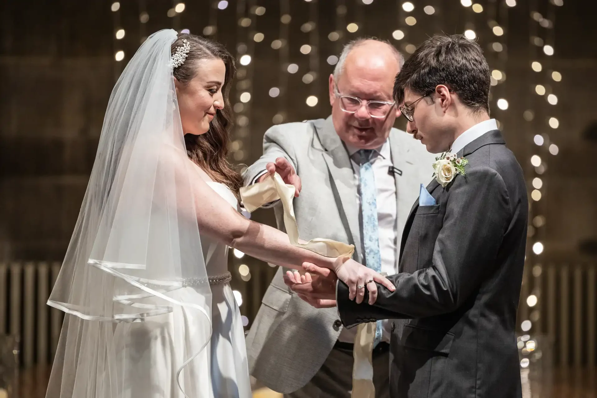 A couple stands facing each other during a wedding ceremony, holding hands. A man officiates, wrapping a ribbon around their hands, with twinkling lights in the background.