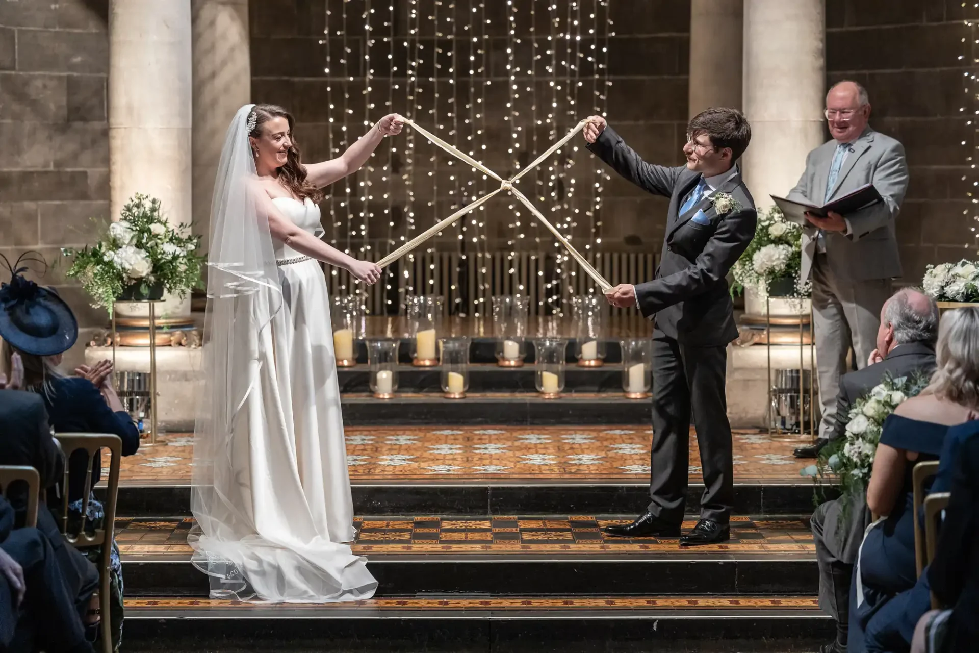 Bride and groom hold long sticks forming an X during a wedding ceremony in a church setting, with guests seated nearby and a man standing behind them holding papers.