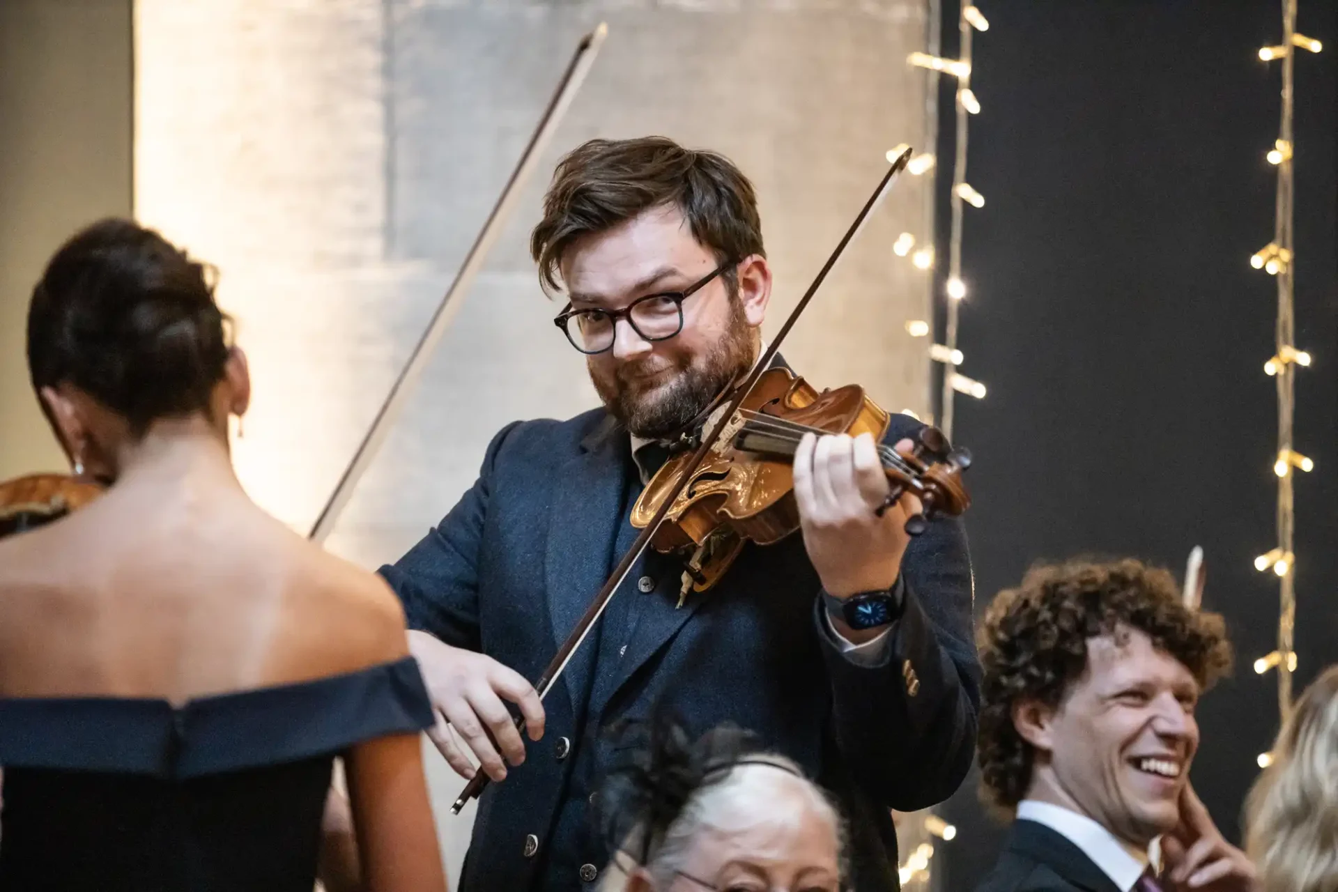 A man plays a violin at an event, surrounded by other musicians. He wears glasses and a dark suit. String lights are visible in the background.