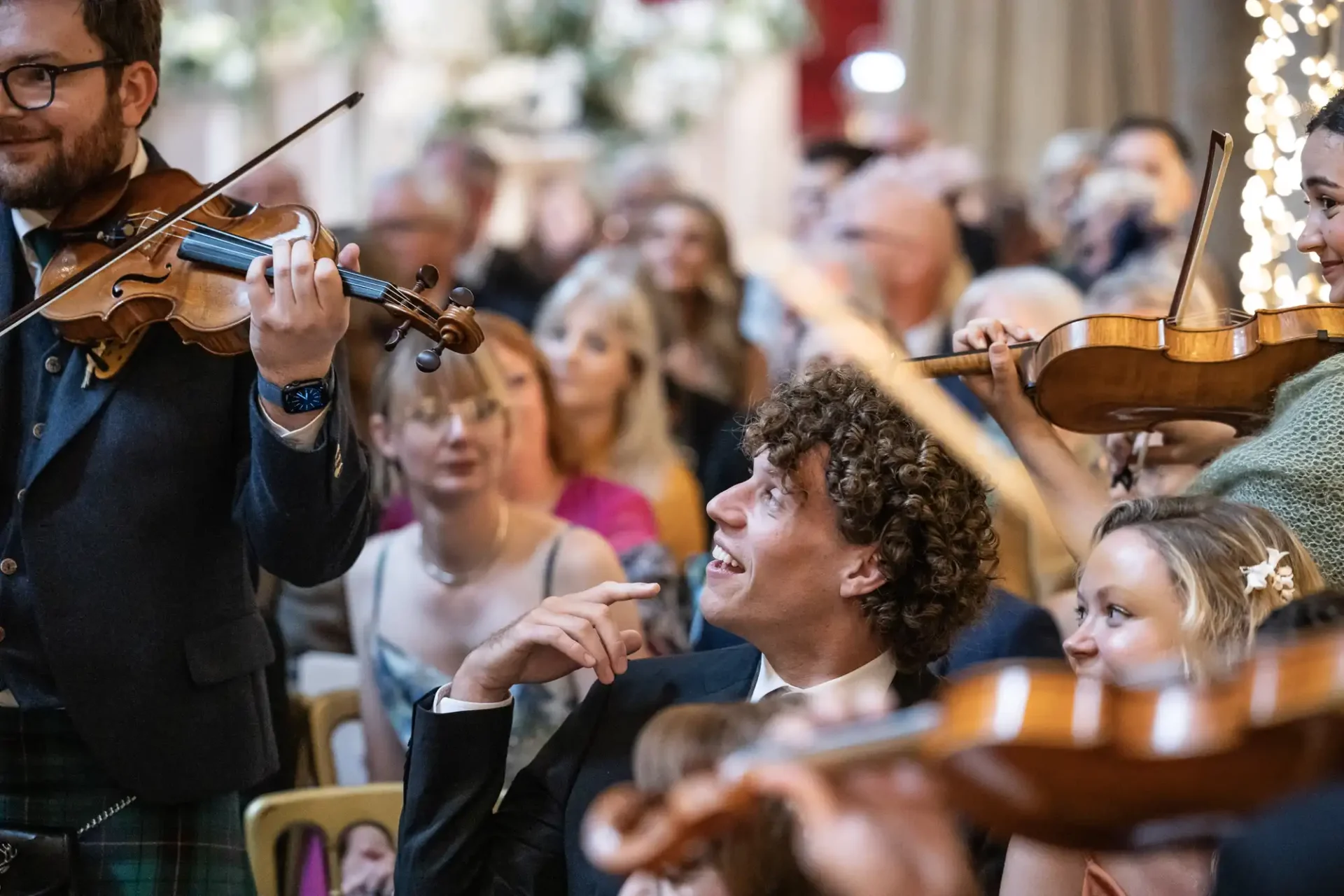 A group of musicians play string instruments at an indoor event, while a man in the audience looks up at them with interest.