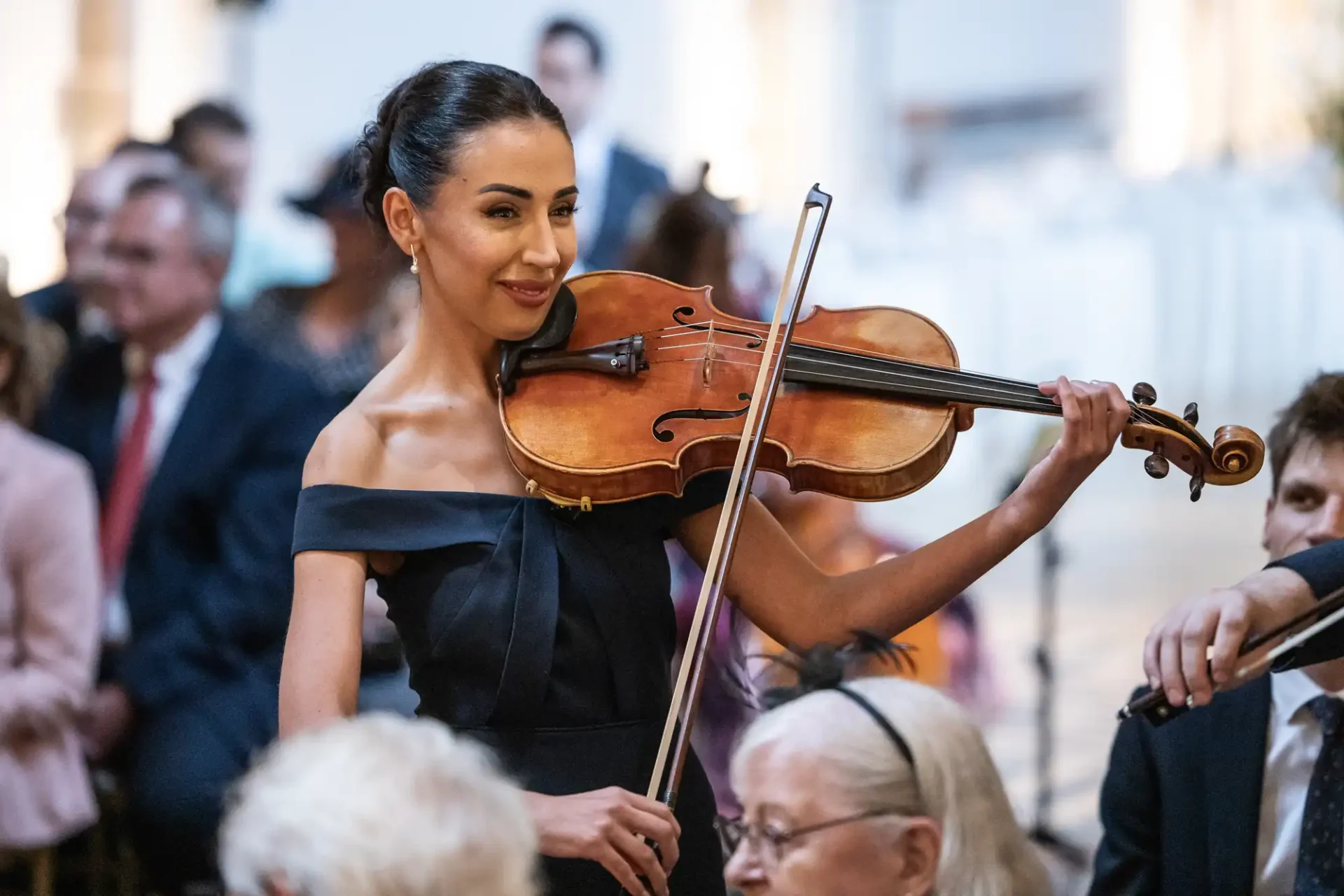 A woman in a black dress plays a violin during a formal event, surrounded by seated guests.