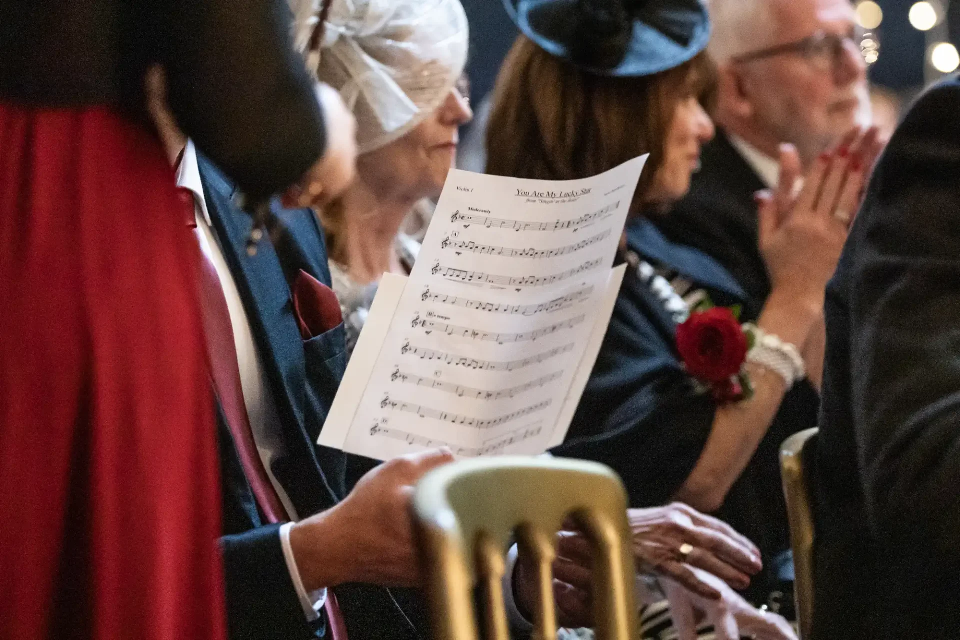 A person sitting in a chair holds sheet music titled "You Are My Sunshine" in a venue with several other seated people nearby.