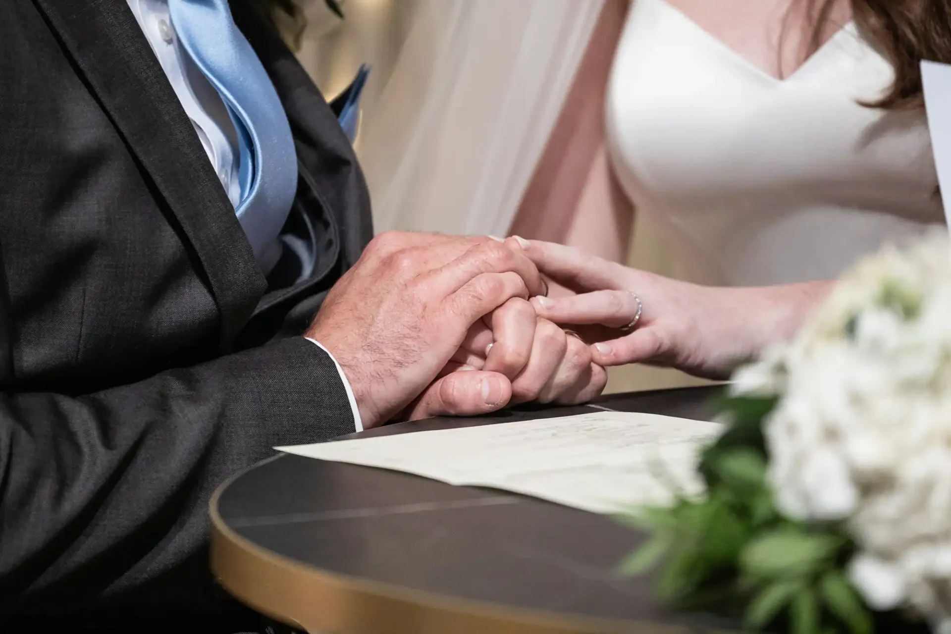 A couple exchanges rings during a wedding ceremony, with their hands placed on a table next to a bouquet of flowers and a document.