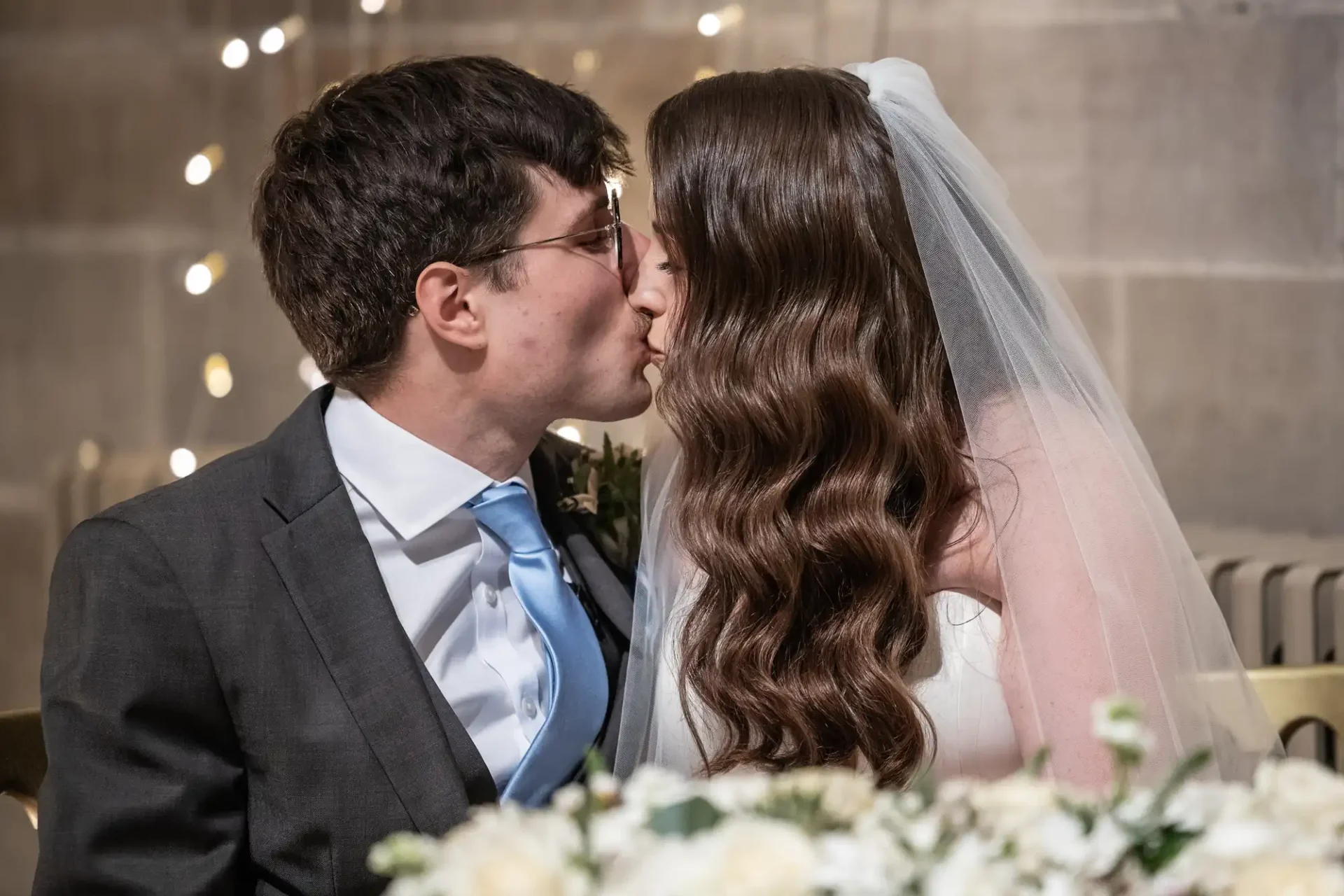 A bride and groom kiss in a wedding setting, with the bride in a white veil and the groom in a suit with a blue tie.