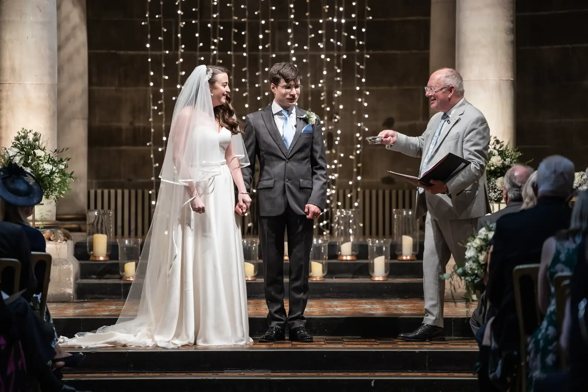 A bride and groom stand hand in hand during a wedding ceremony, officiated by a man holding a book. They are surrounded by candles and string lights. Guests are seated in front.