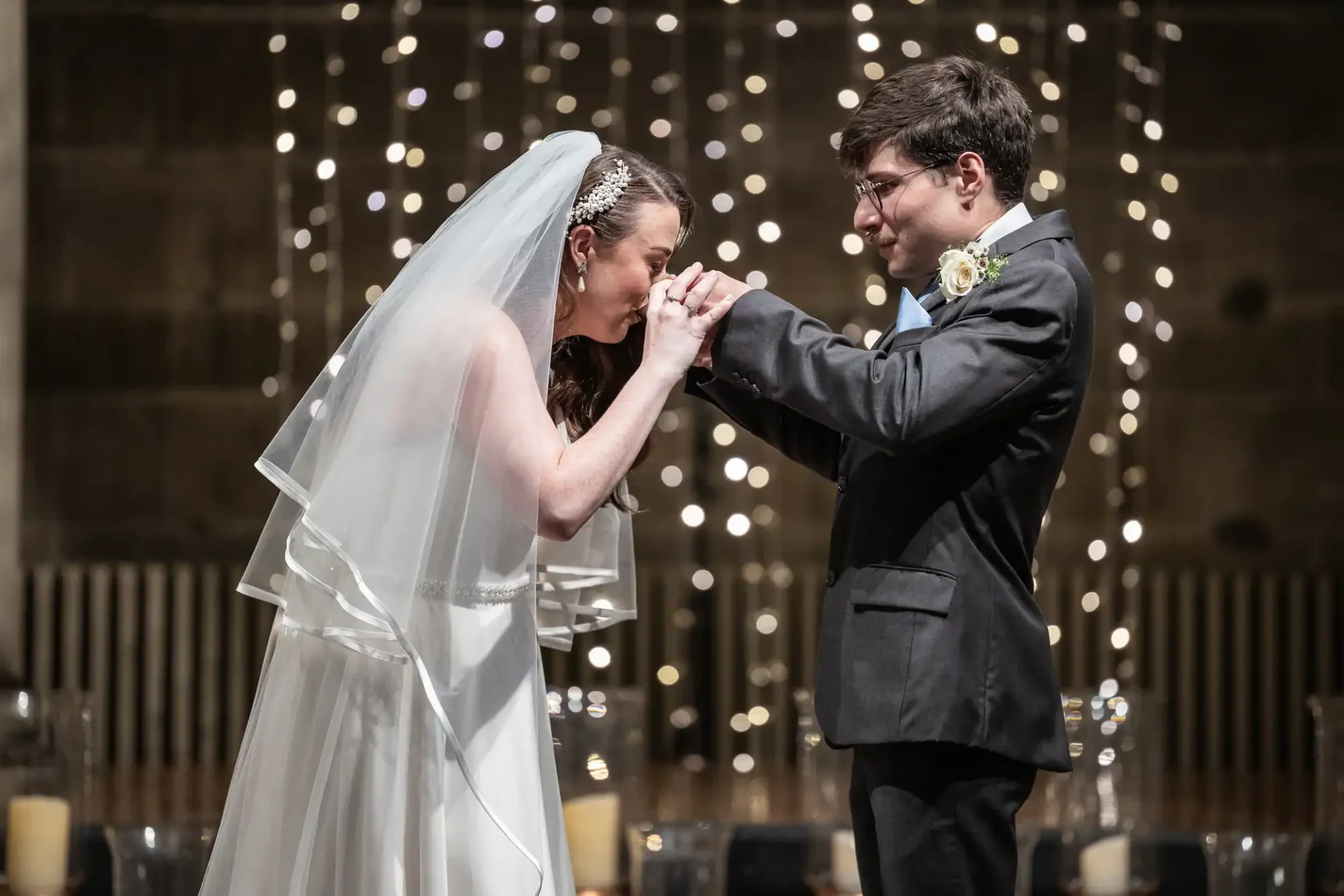 Bride and groom stand under string lights; the bride delicately kisses the groom's hands, while both are smiling at each other.