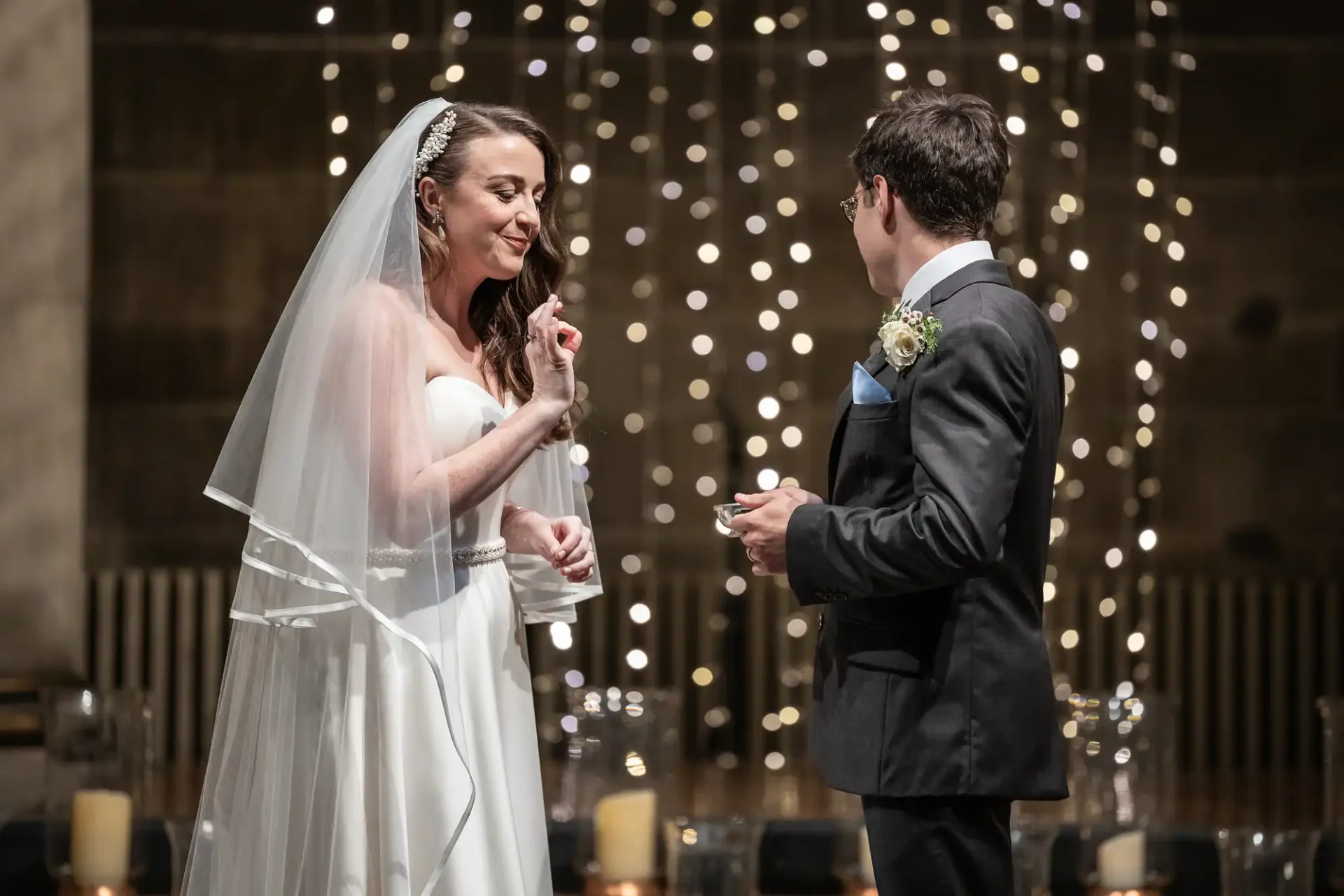 A bride and groom exchange vows during a wedding ceremony, standing in front of a backdrop with string lights and candles.