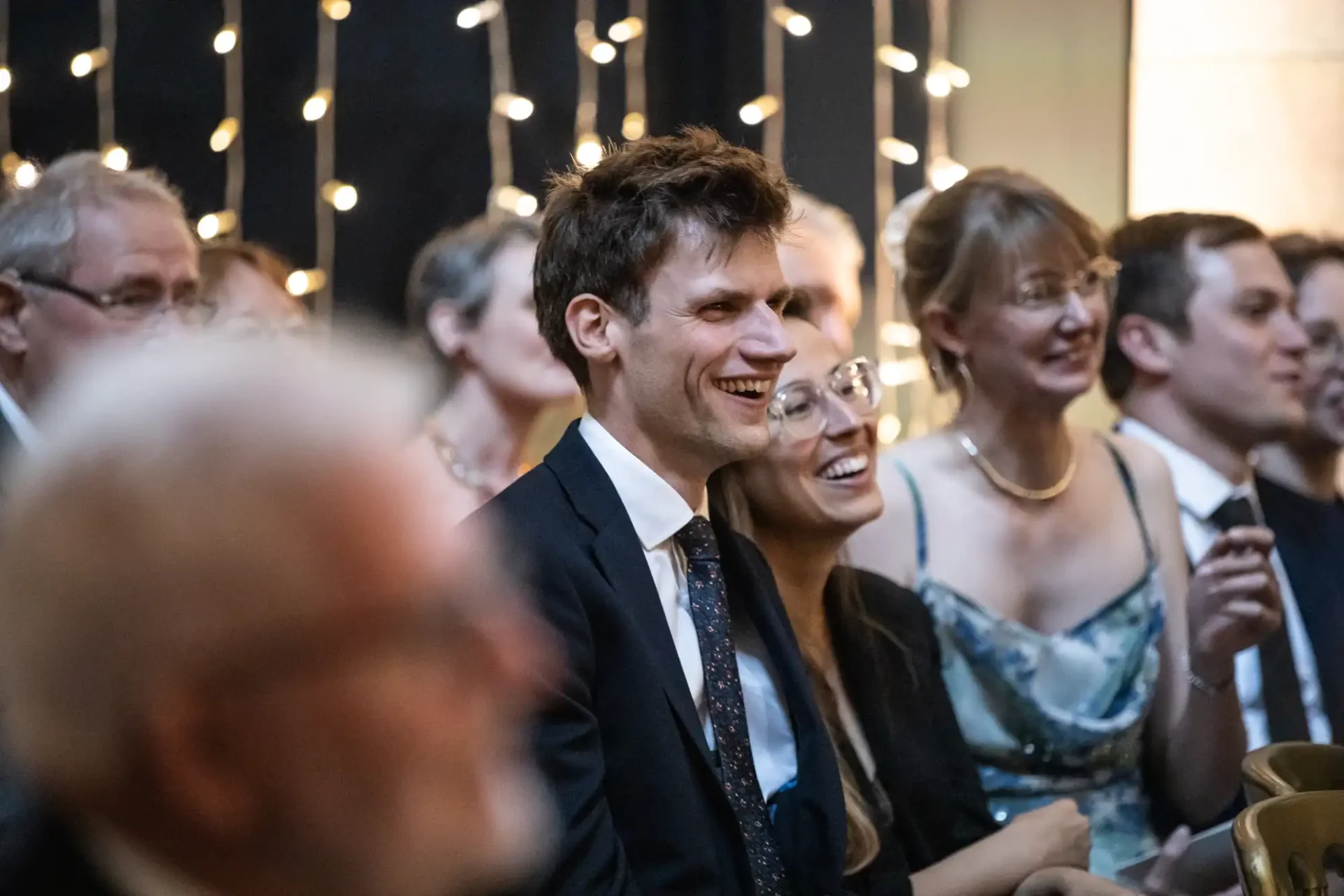 People seated and smiling at an event, with string lights in the background.