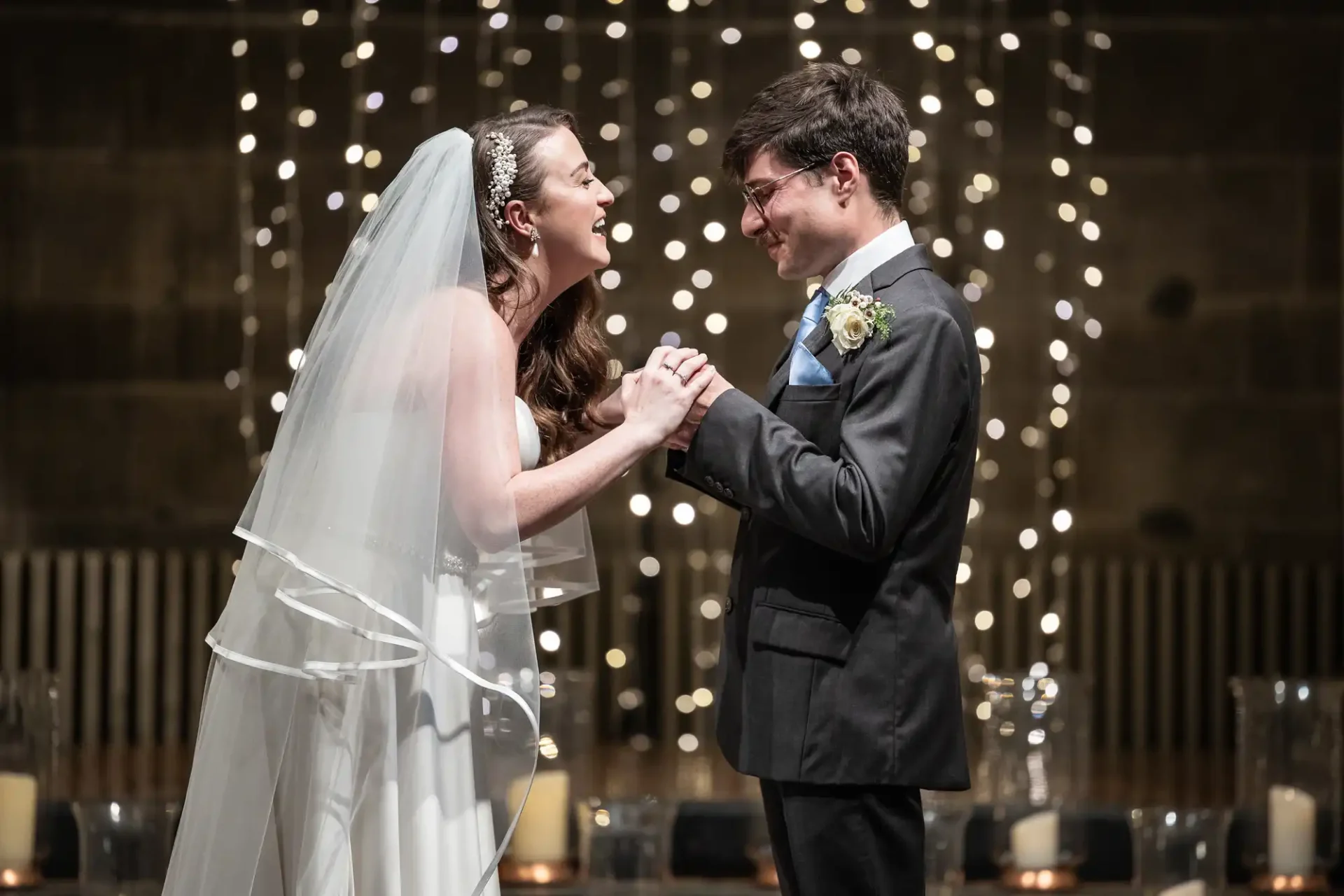 A bride and groom smile and hold hands in front of a backdrop with string lights.