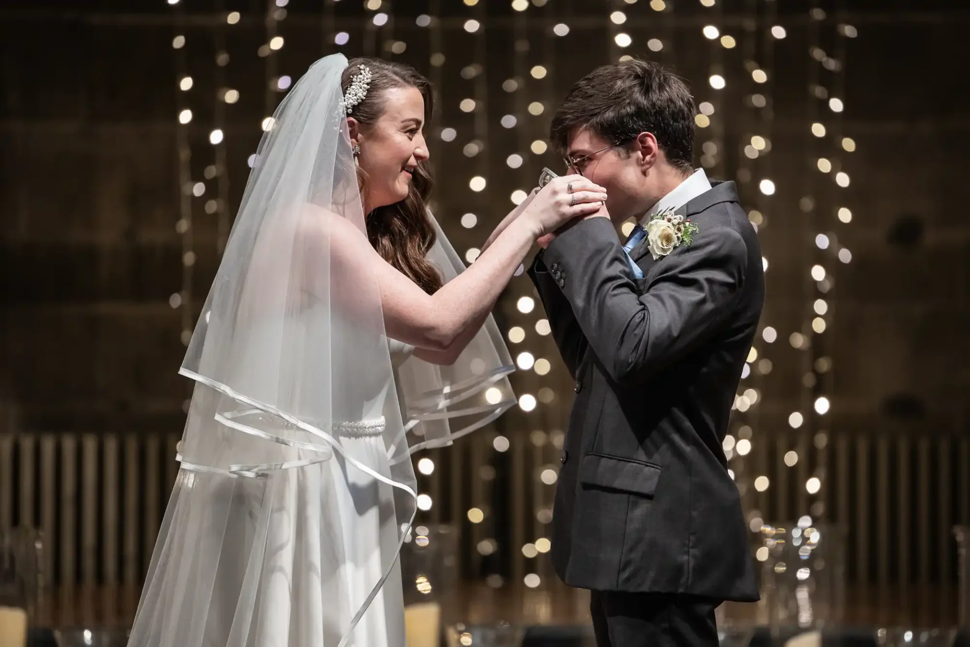 Bride and groom smiling, holding hands, under twinkling lights during a wedding ceremony.