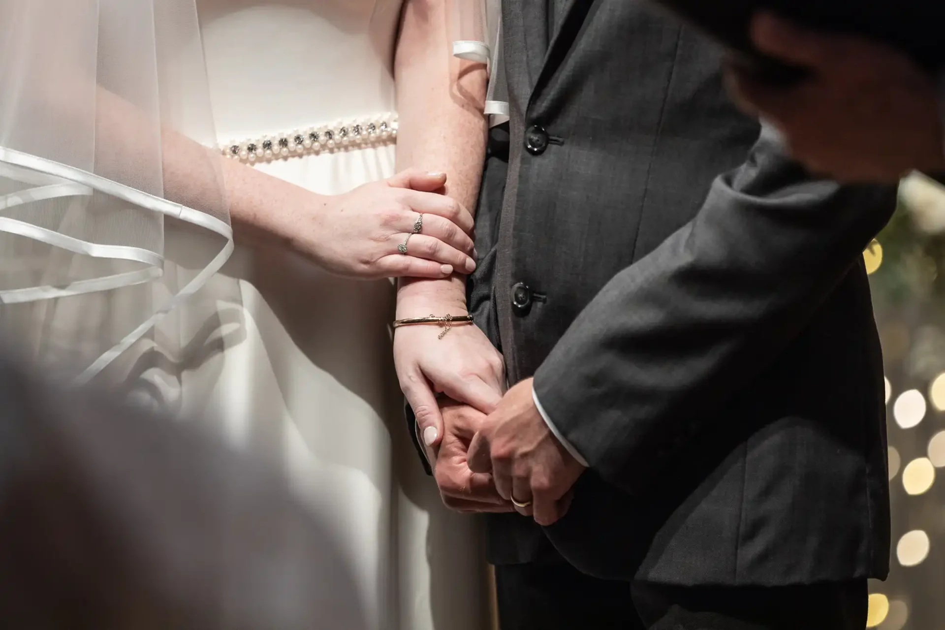 Bride and groom holding hands during wedding ceremony, dressed in wedding attire with blurred background.