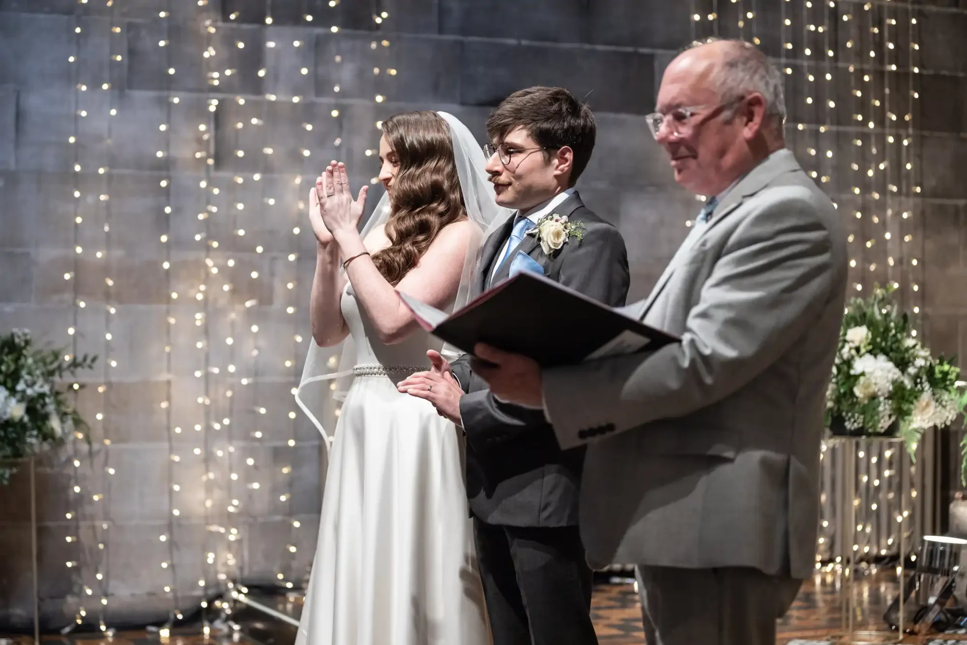 A bride and groom stand next to an officiant holding a book during a wedding ceremony, with string lights and floral arrangements in the background.