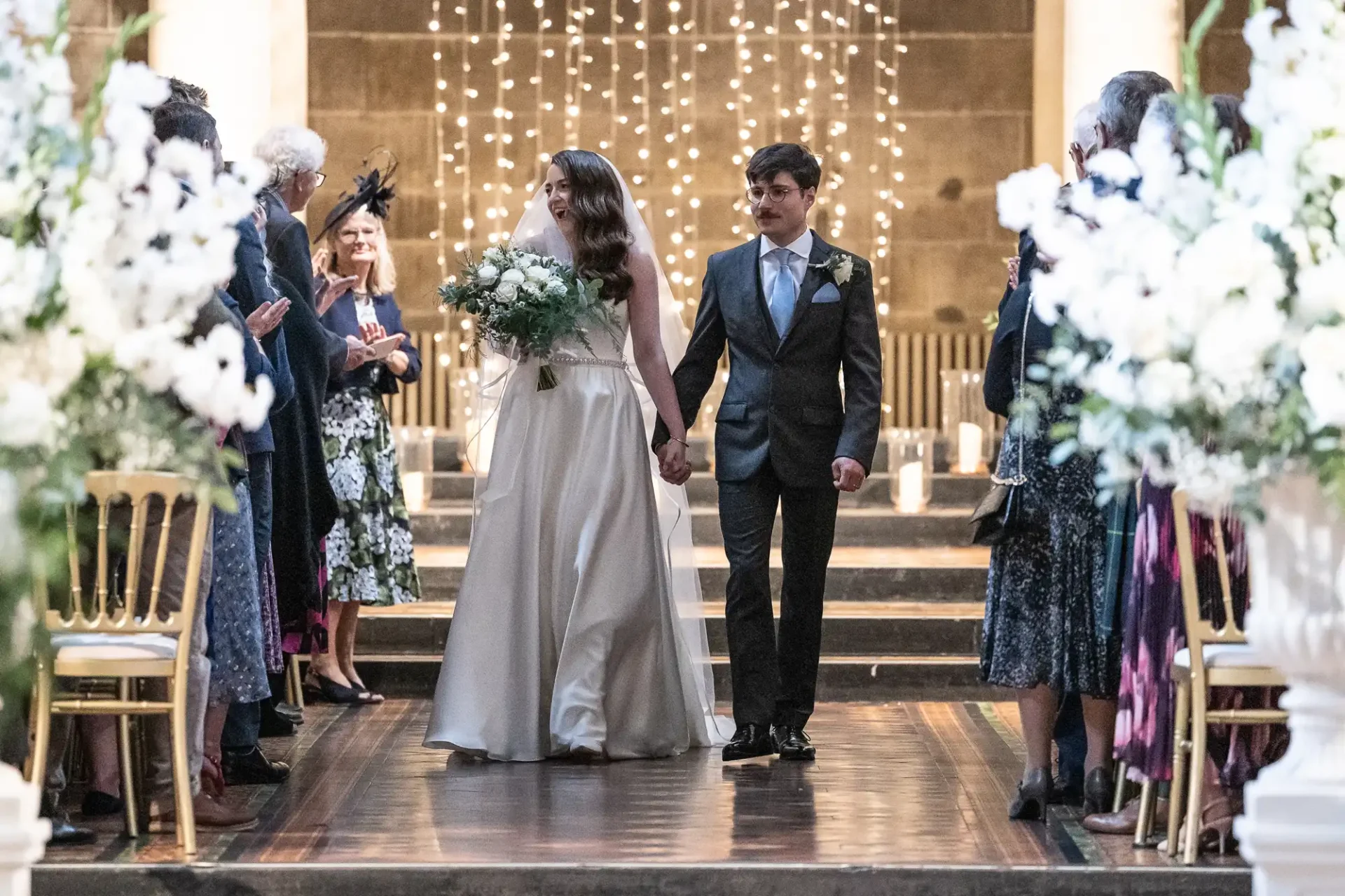 A bride and groom walk down the aisle at a wedding ceremony, holding hands. They are surrounded by guests and white floral decorations. String lights are visible in the background.