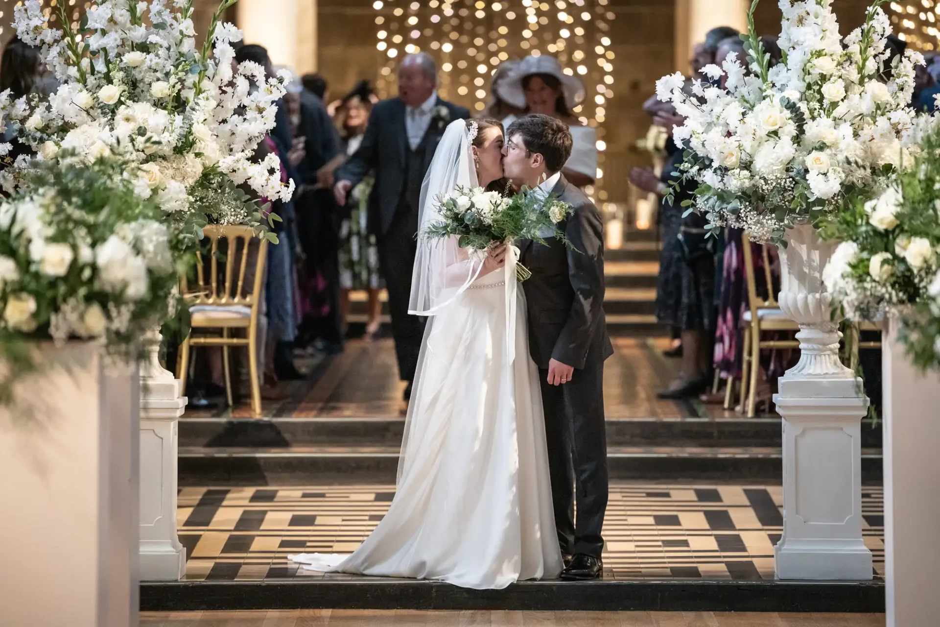 A bride and groom kiss at the altar, surrounded by white flowers and guests seated in the background.