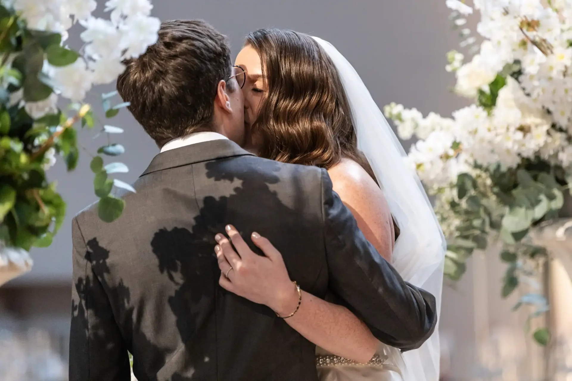 A bride and groom share a kiss at their wedding, surrounded by white flowers and greenery.