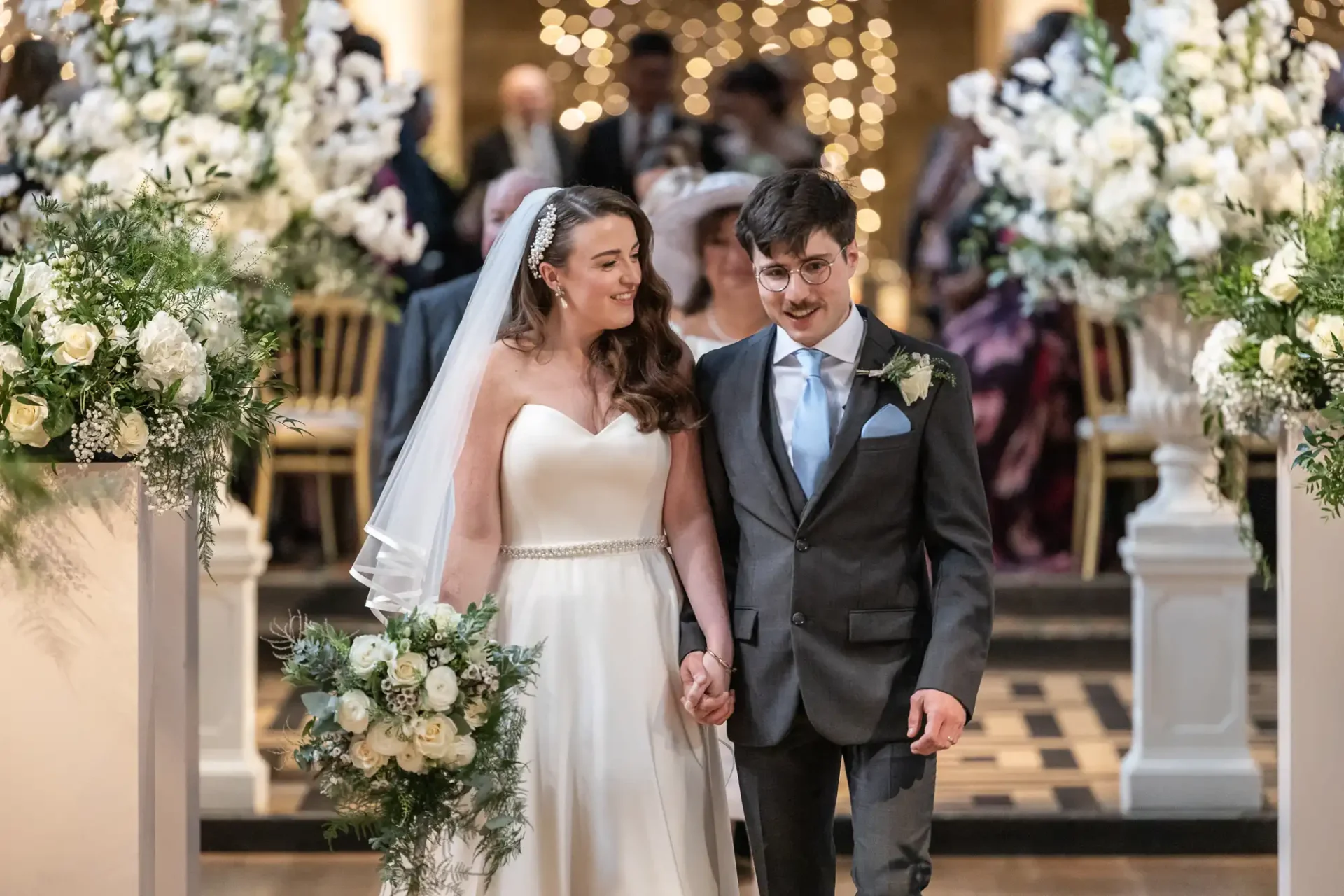 A bride and groom walk down the aisle, holding hands. The bride wears a white dress and veil, carrying a bouquet. The groom is in a suit with a tie. White floral arrangements are visible.