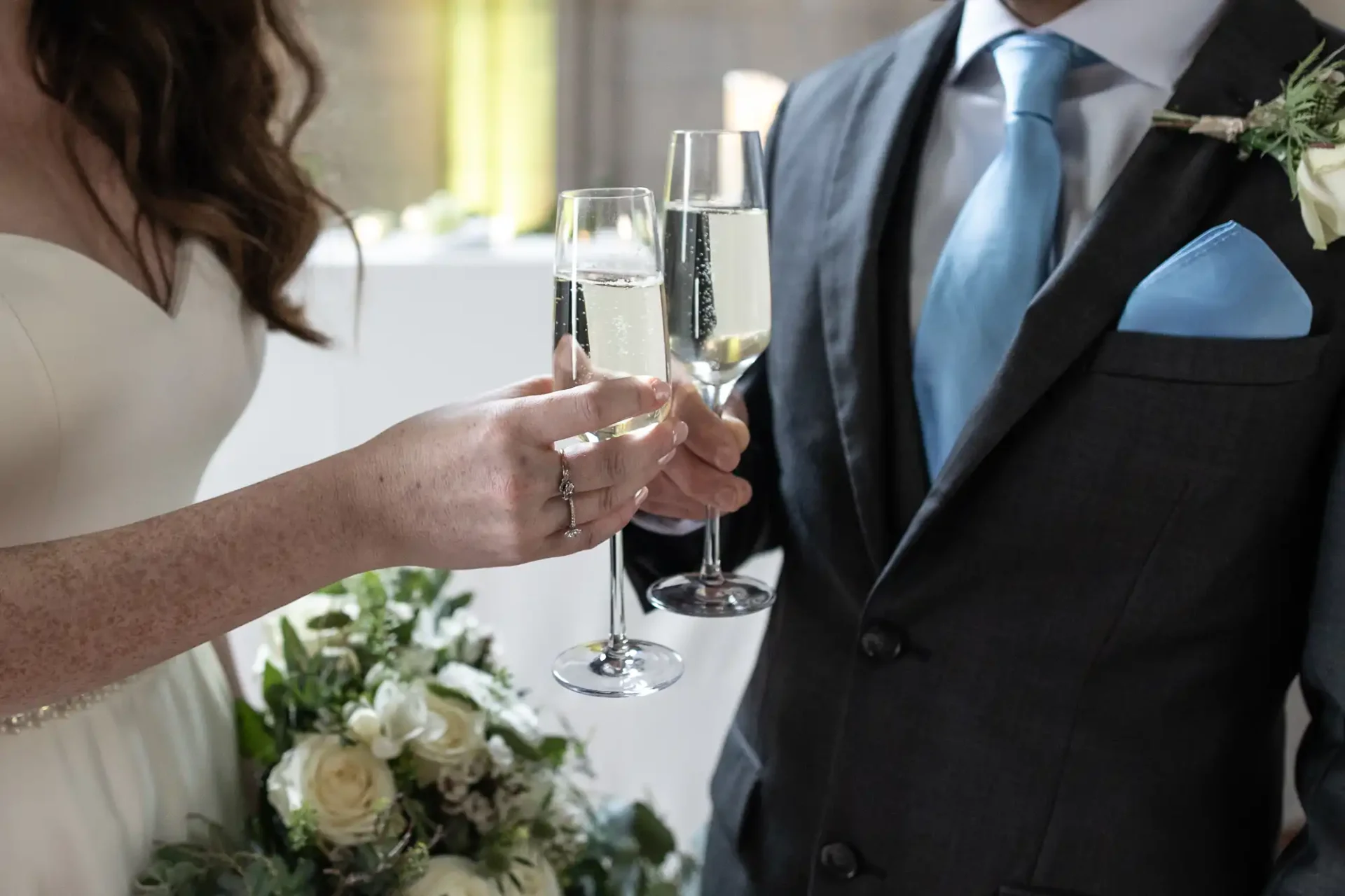 Bride and groom toast with champagne glasses, holding a bouquet, in formal attire.