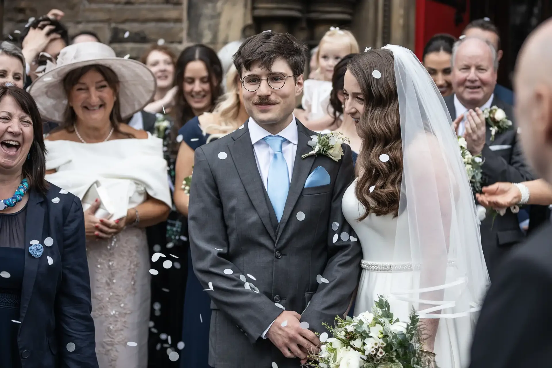 A bride and groom stand together outdoors, surrounded by guests, with confetti falling around them. The groom wears a suit and the bride a white dress and veil, holding a bouquet.