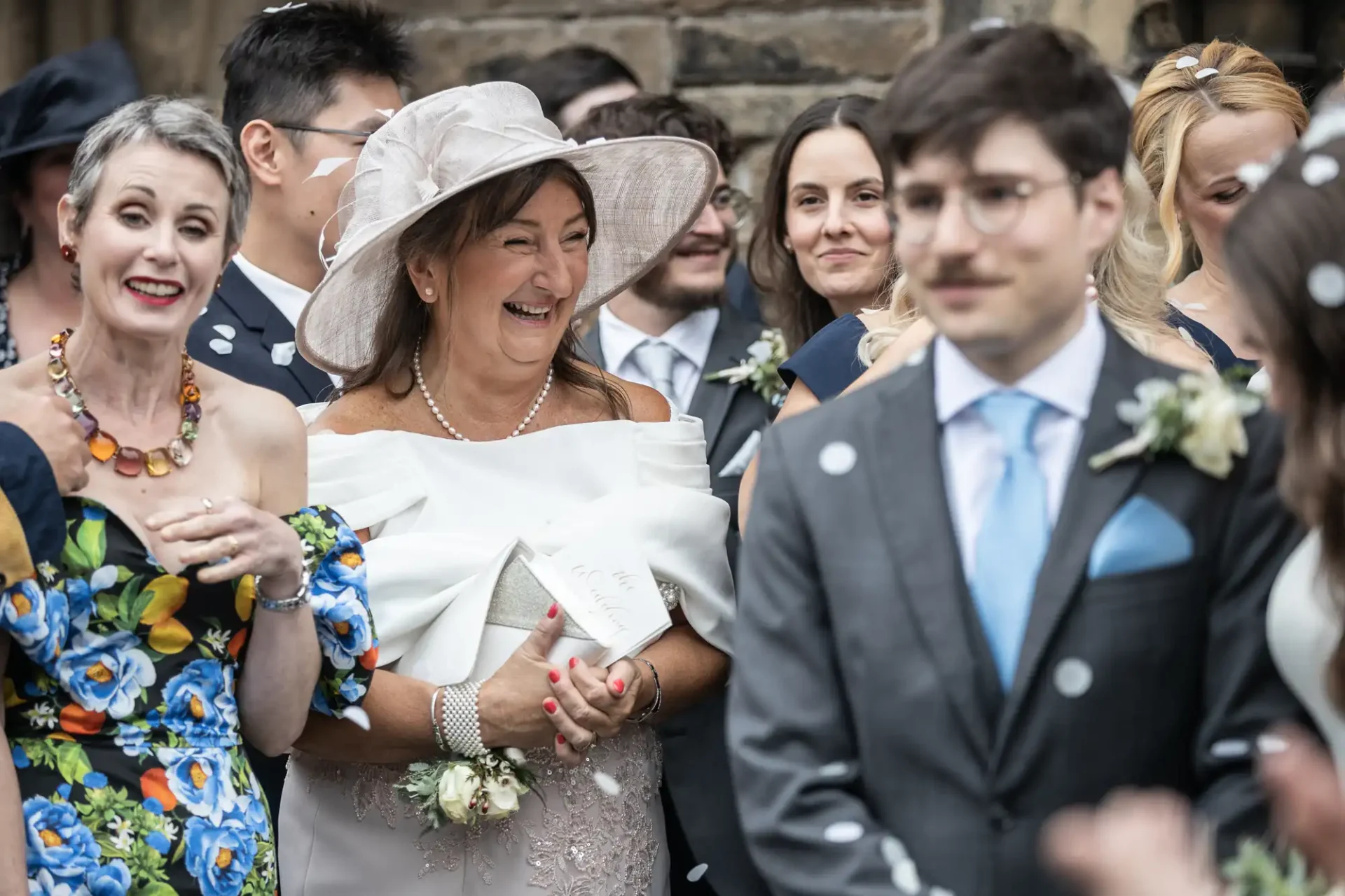 Group of people at a wedding, smiling and dressed formally. A woman in a floral dress and a woman in a large hat are in the foreground. Confetti is being thrown.