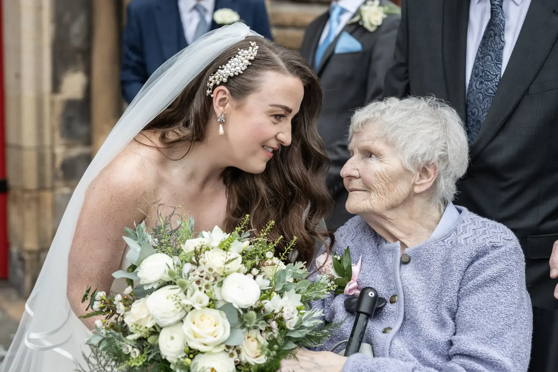 A bride in a white gown and veil holds a bouquet, leaning towards an elderly woman in a lilac cardigan seated in a wheelchair. Both are smiling at each other.