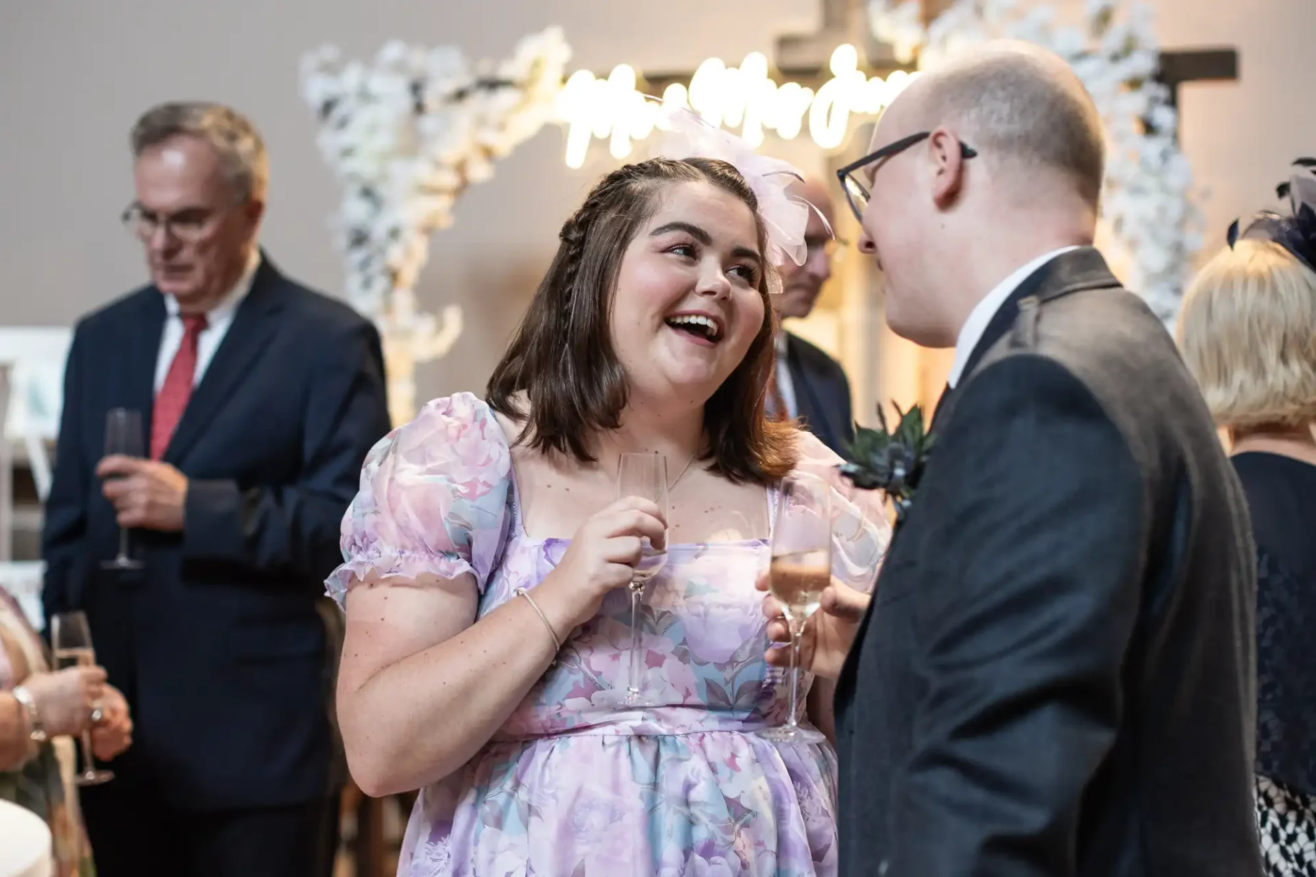 A woman in a floral dress smiling at a man in a suit, both holding drinks. Other people in suits and dresses are in the background, mingling at an indoor event.