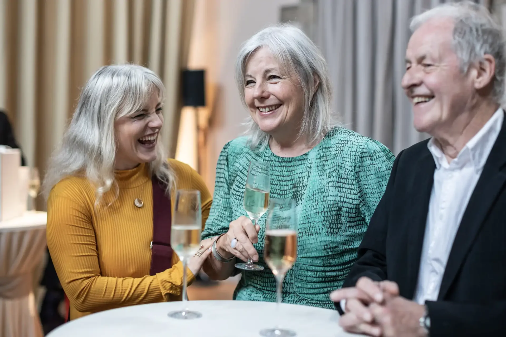 Three people smiling and holding champagne glasses at a table in a warmly lit room.