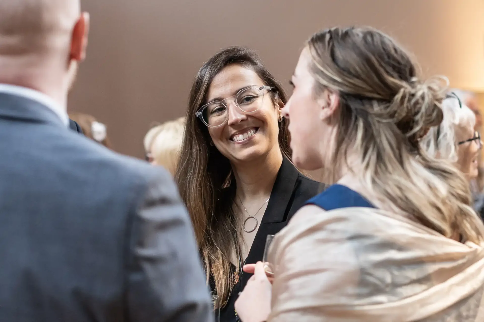 Three people engaged in conversation at an indoor event. The woman in glasses is smiling while listening to another woman who has her back to the camera.