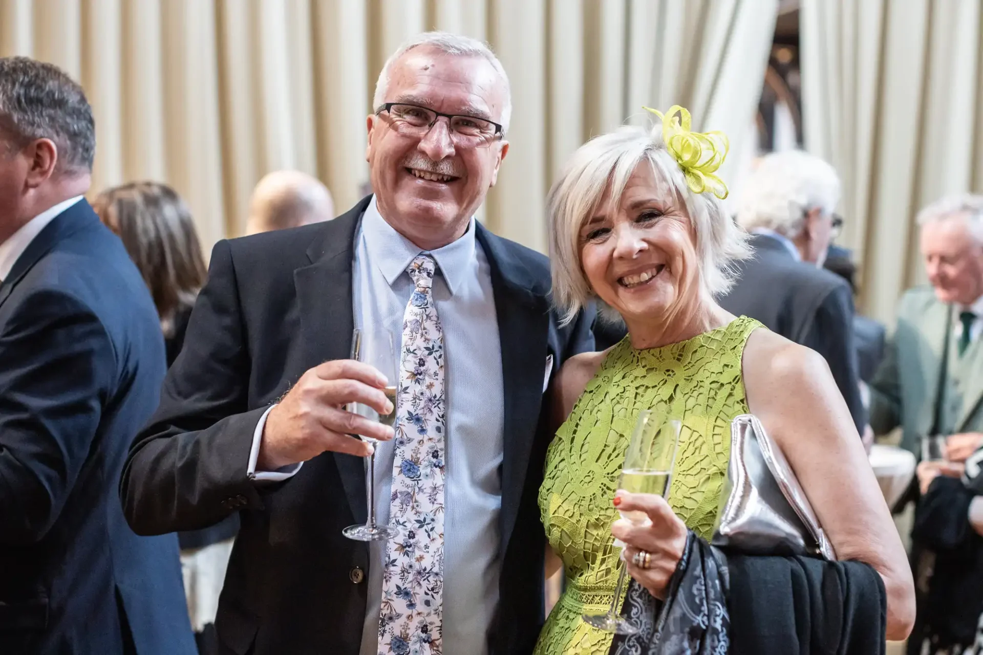 An older couple at an event, smiling and holding glasses. The man wears a suit and patterned tie; the woman in a yellow dress with a floral headpiece carries a purse. People in the background.