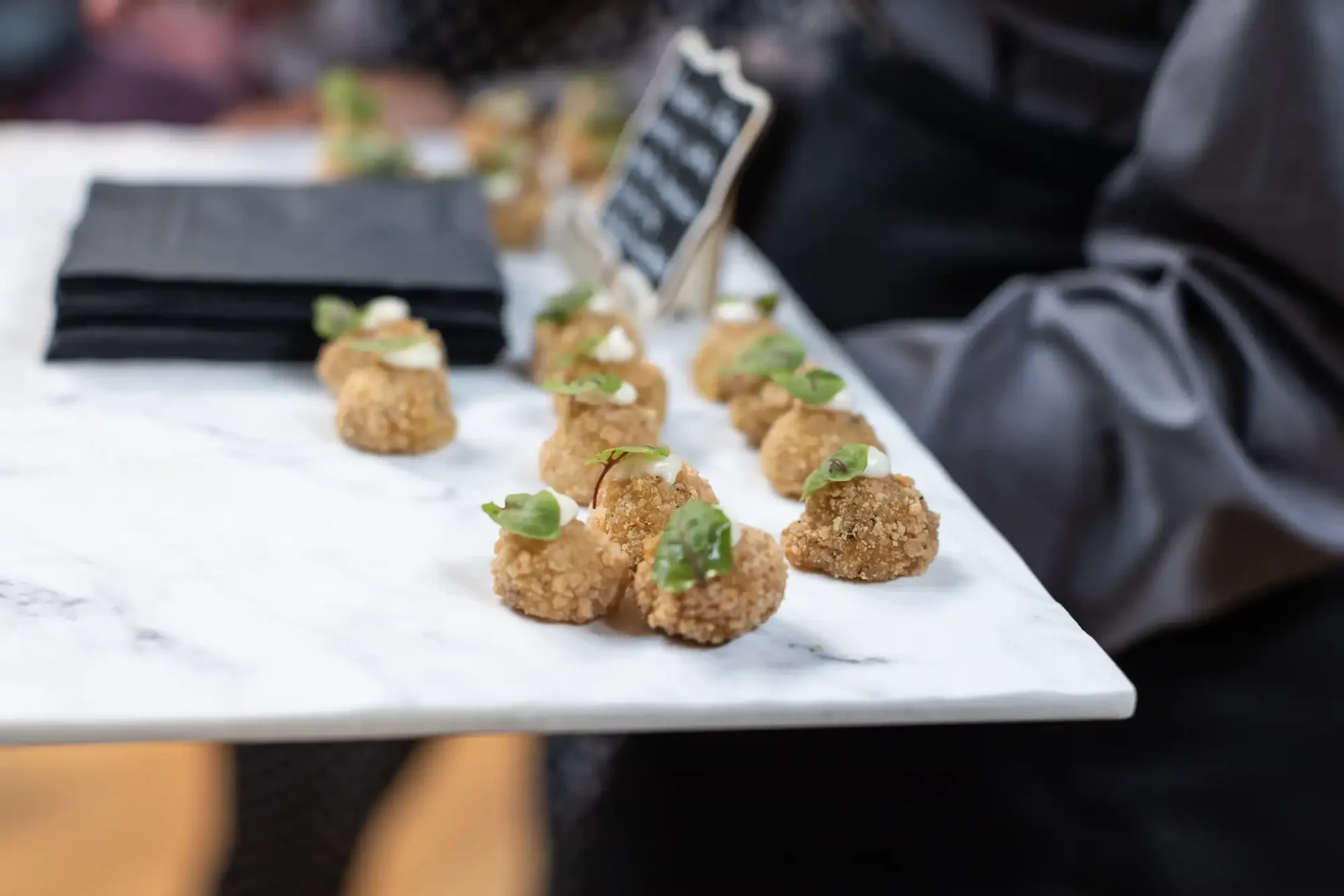 Close-up of a marble tray with bite-sized appetizers, likely croquettes, garnished with small greens. A person in a gray shirt holds the tray, and a menu card is visible in the background.