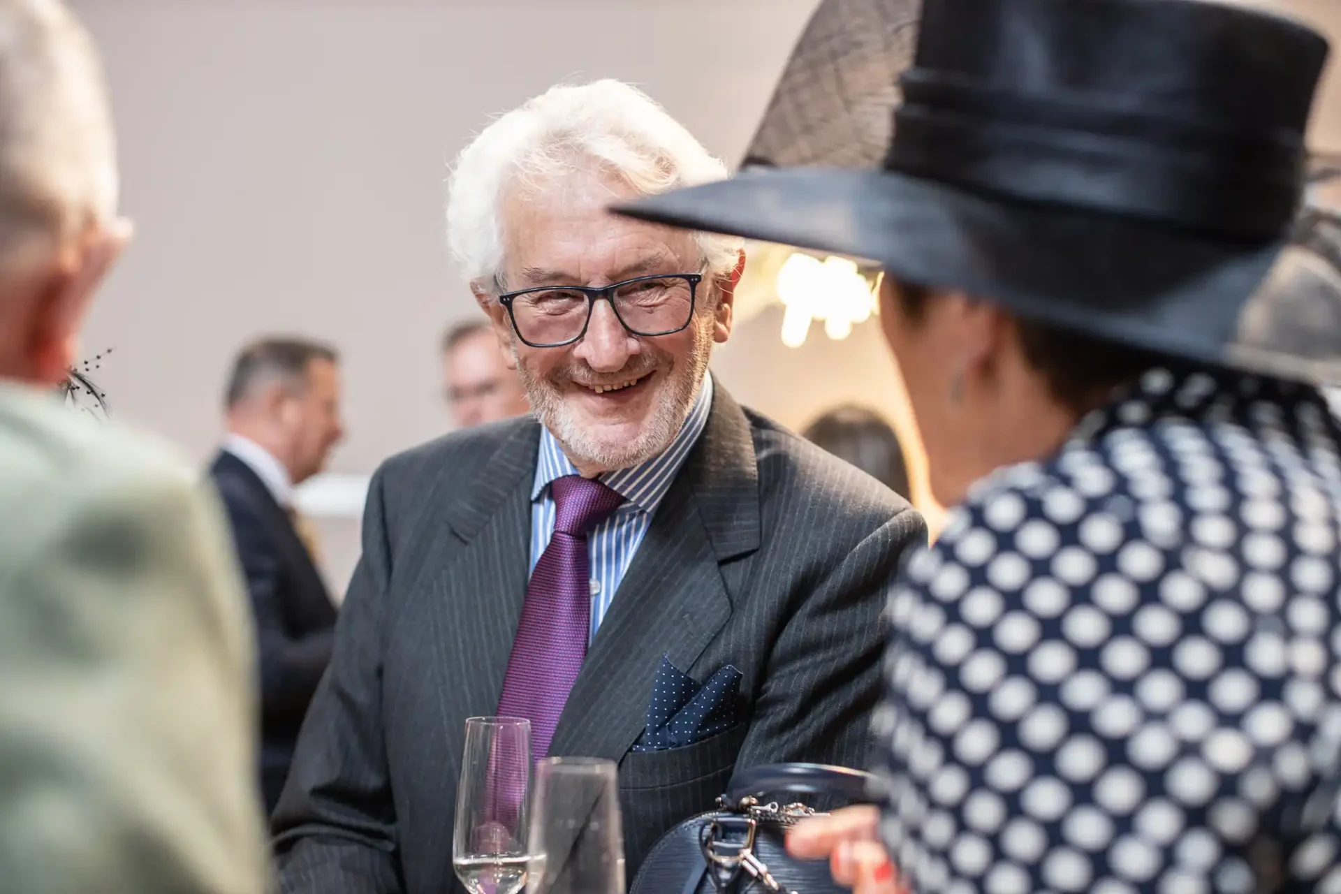 An elderly man in a suit and tie smiles while conversing with a woman in a polka dot outfit and large hat at an event, with wine glasses on the table.