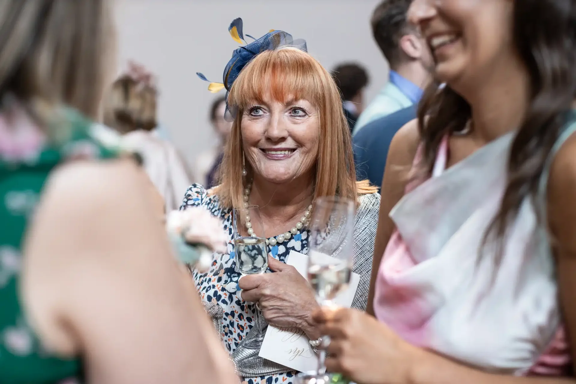 A woman with red hair wearing a fascinator is smiling while holding a glass at a social gathering, surrounded by people.