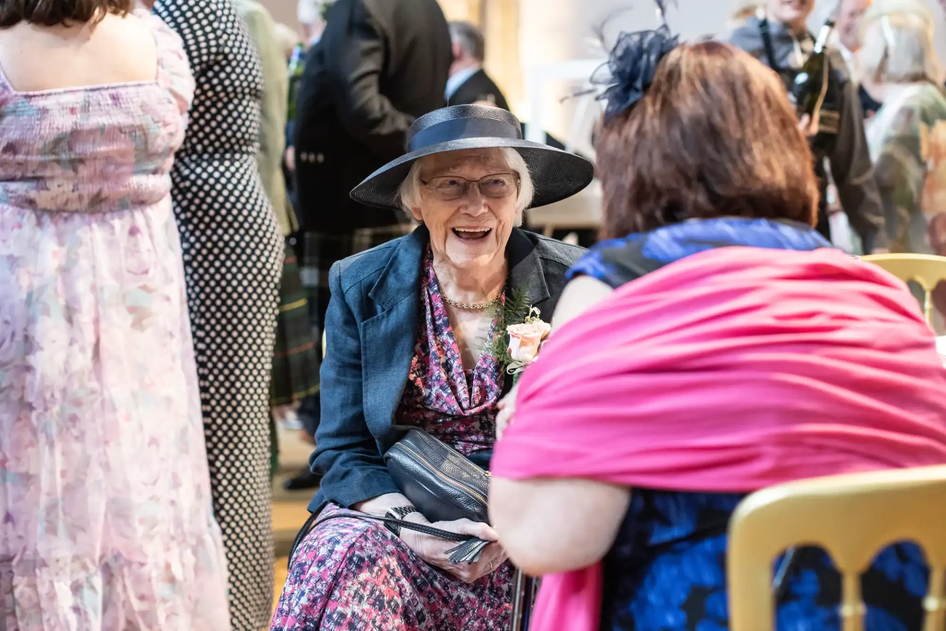 Two women seated, facing each other at an indoor event. One wears a wide-brimmed hat and floral dress. The other wears a pink shawl and blue dress. People and chairs are visible in the background.